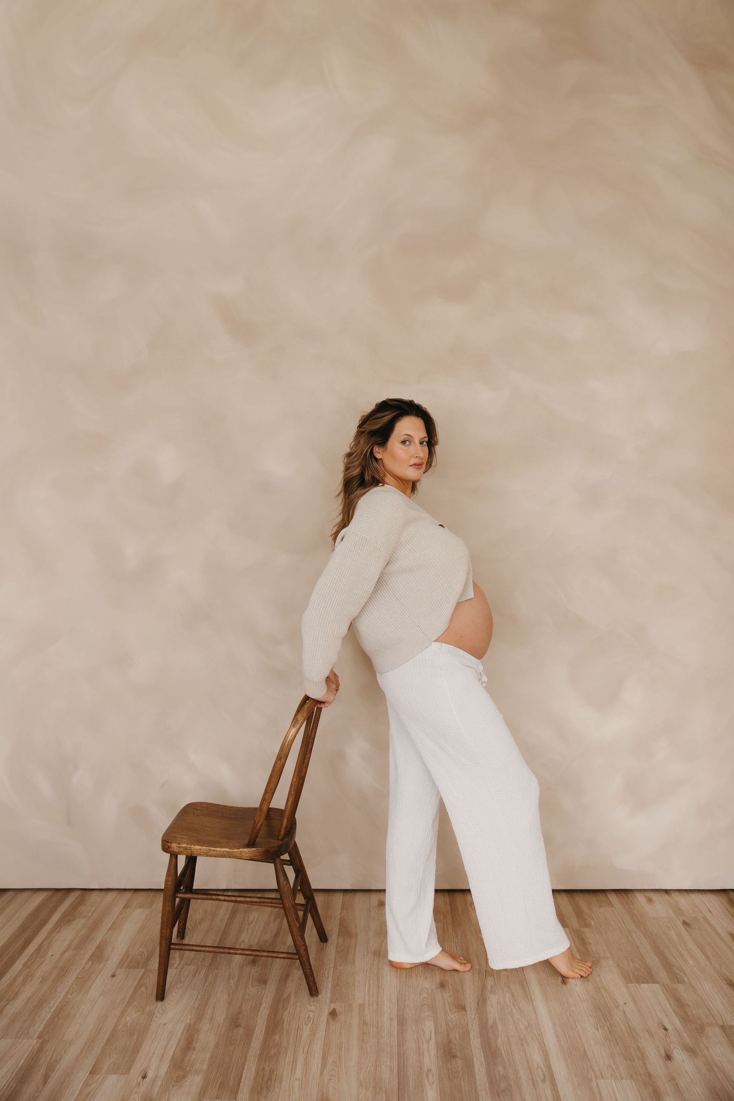 Pregnant woman stands beside wooden chair against beige wall in white pants and fitted top.