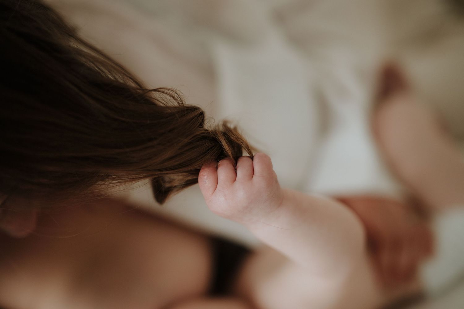 A baby's tiny hand gently grasping a mother's long brown hair while lying on a white bed.