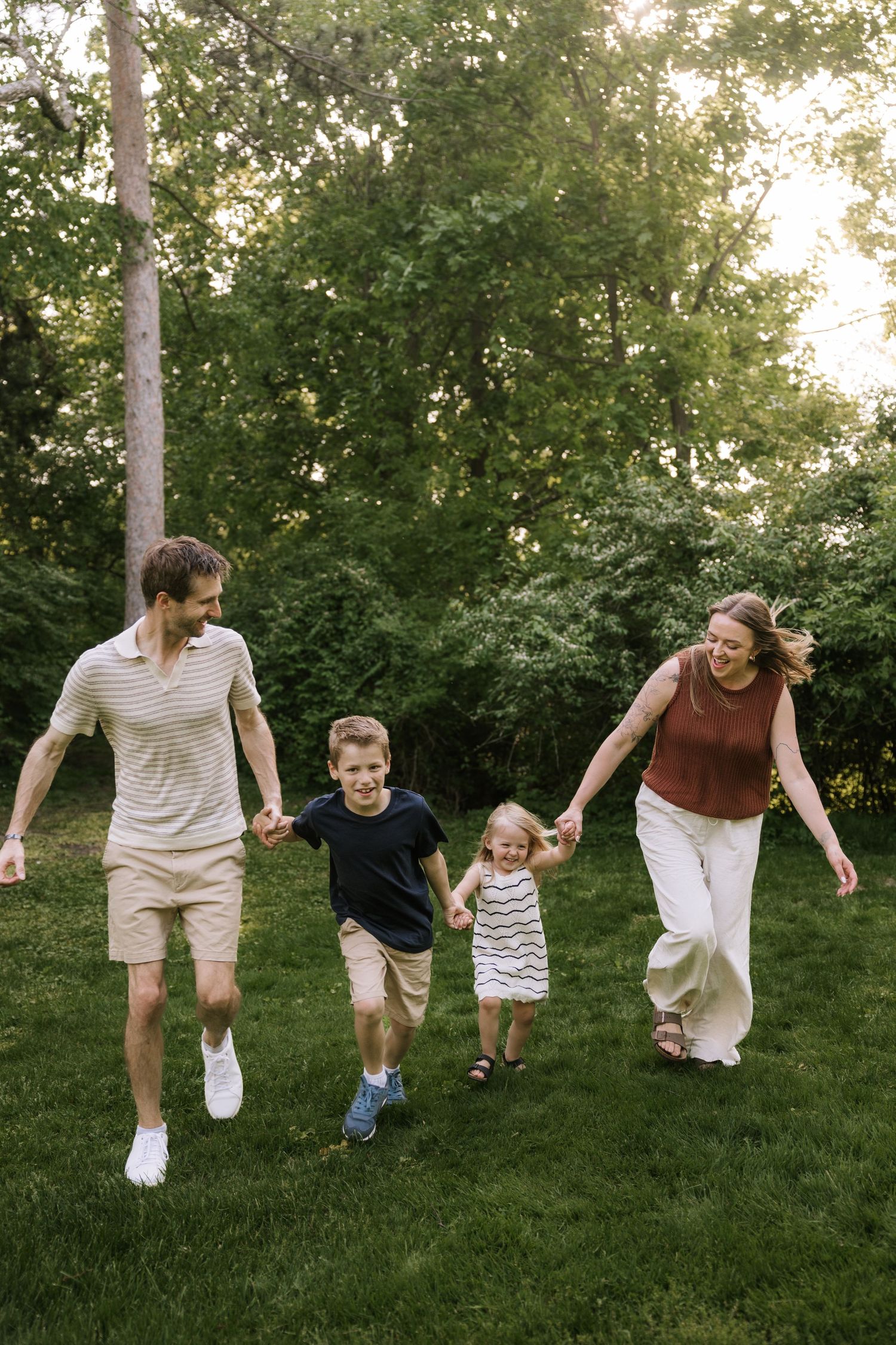 A joyful family of four holds hands while walking through a lush green backyard on a sunny day.