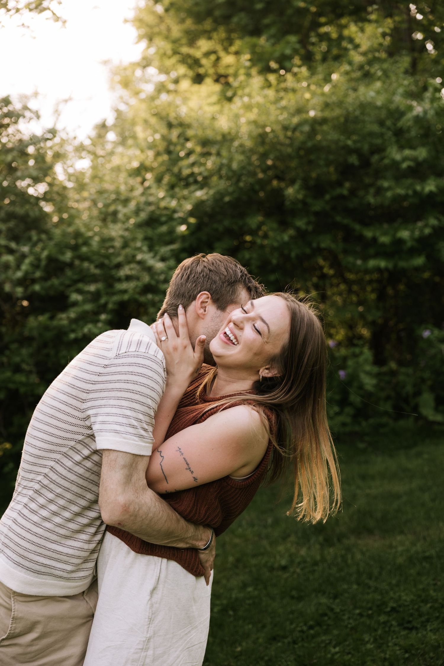 A couple shares a romantic embrace outdoors, with the man kissing the laughing woman on the cheek in golden light.