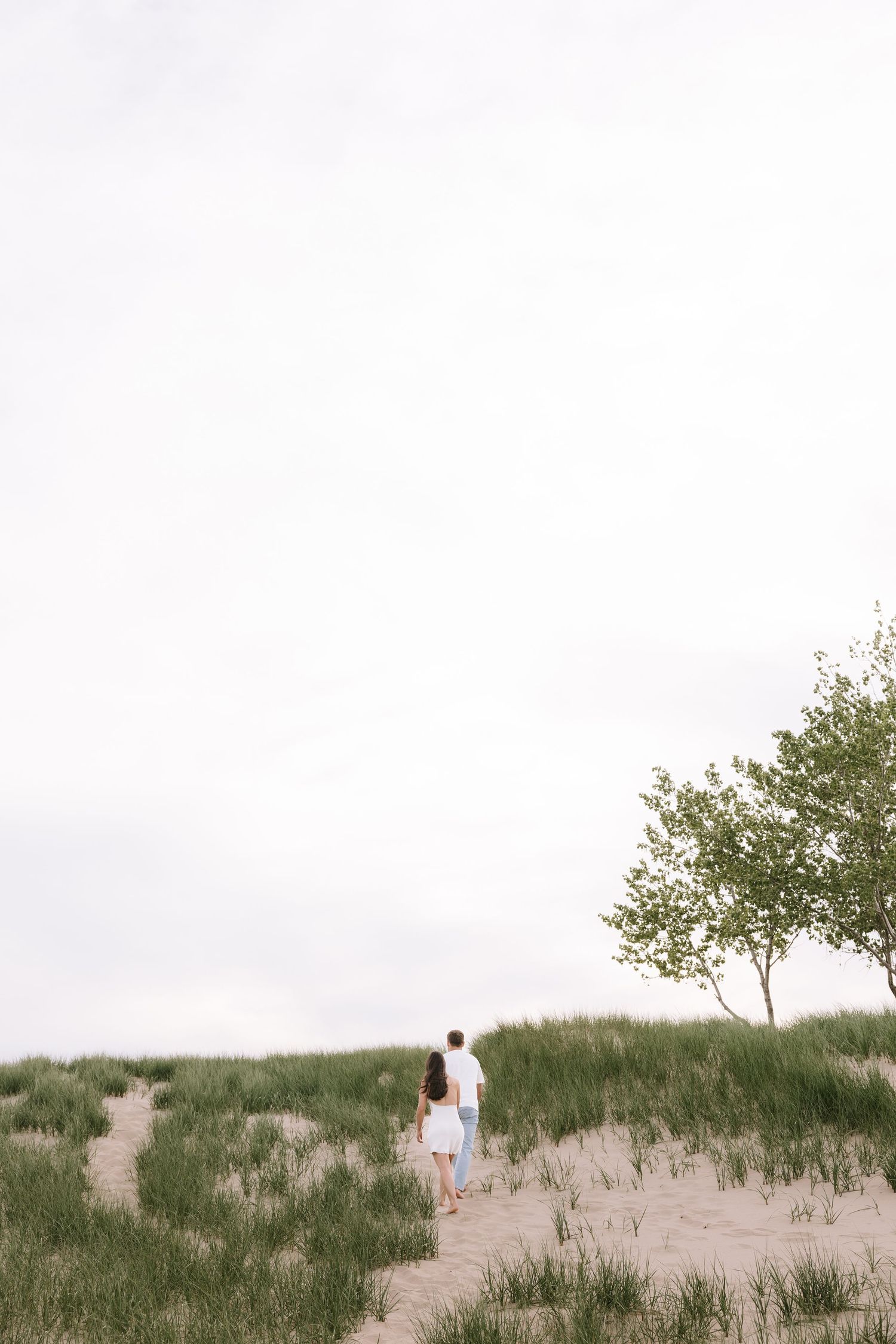 Couple walking hand in hand up sandy dune path with green grass and lone tree under overcast sky.