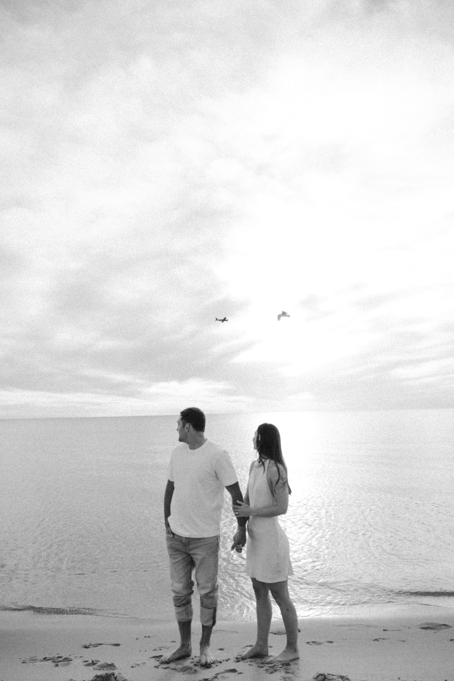Couple holding hands on a beach at sunset in black and white, standing barefoot with birds flying overhead.