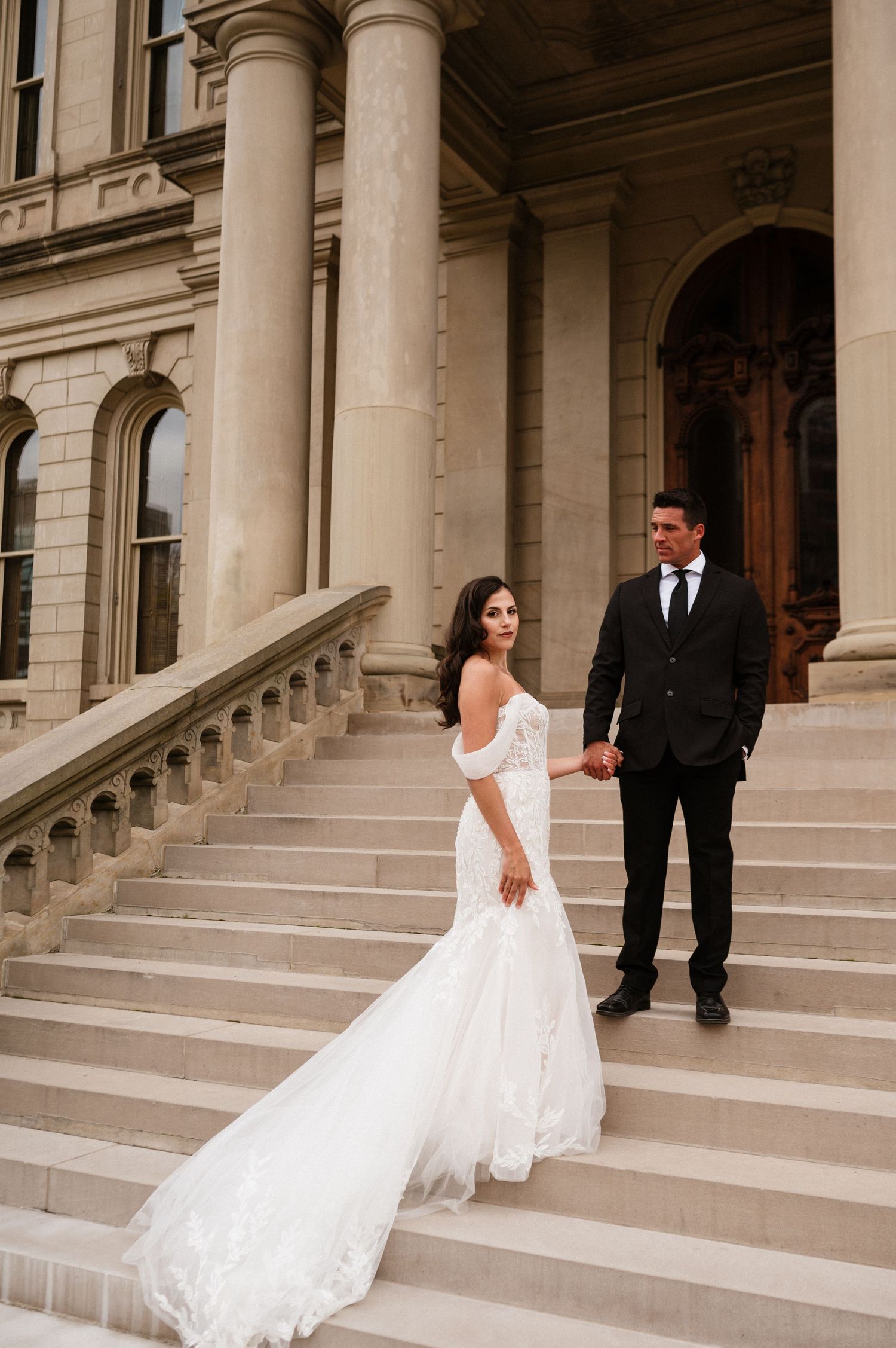 A bride and groom pose on elegant stone steps of a classical building.