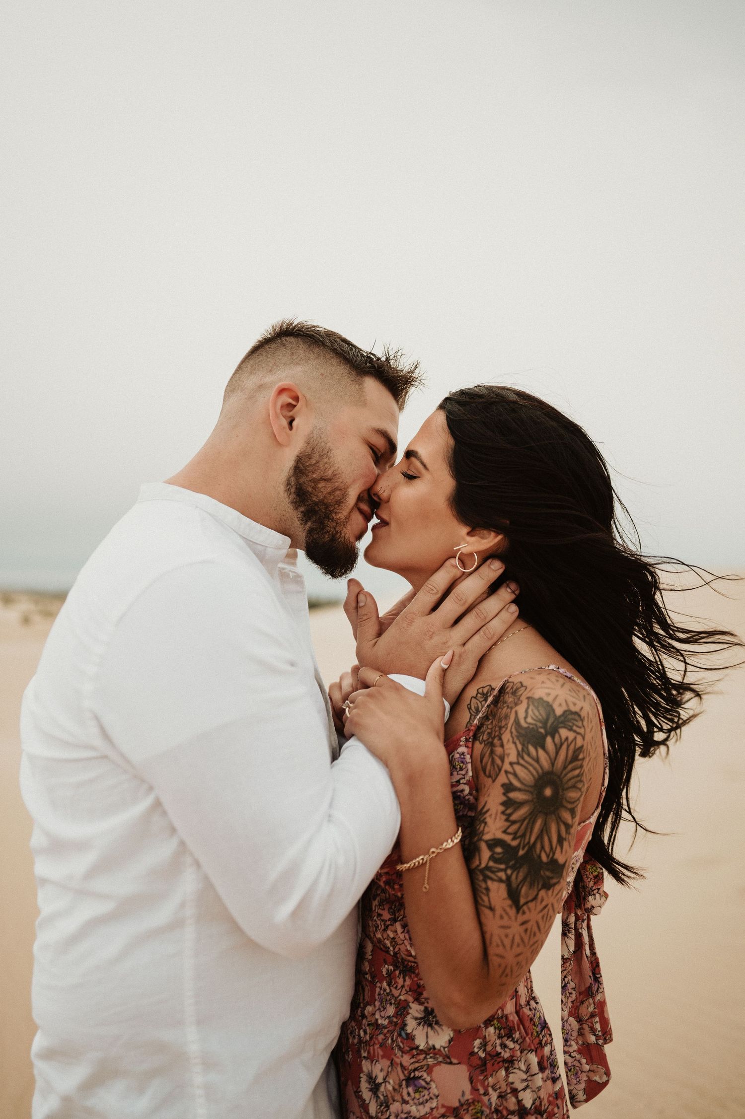 An intimate desert photoshoot captures a couple in white and floral dress embracing against sandy dunes.