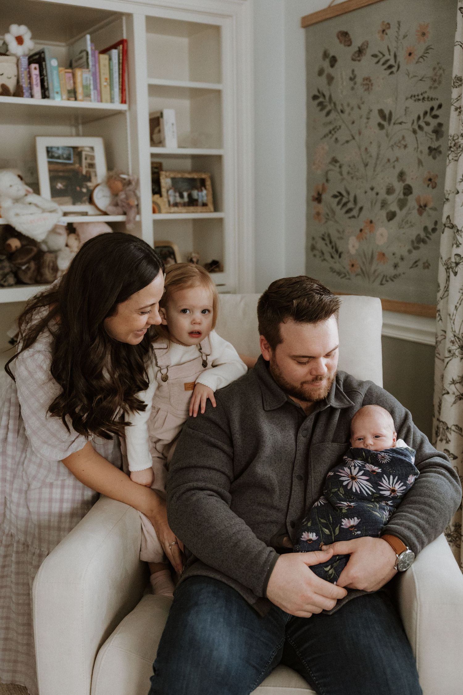 A family relaxes together on a white couch in a cozy living room with floral wallpaper and built-in bookshelves.