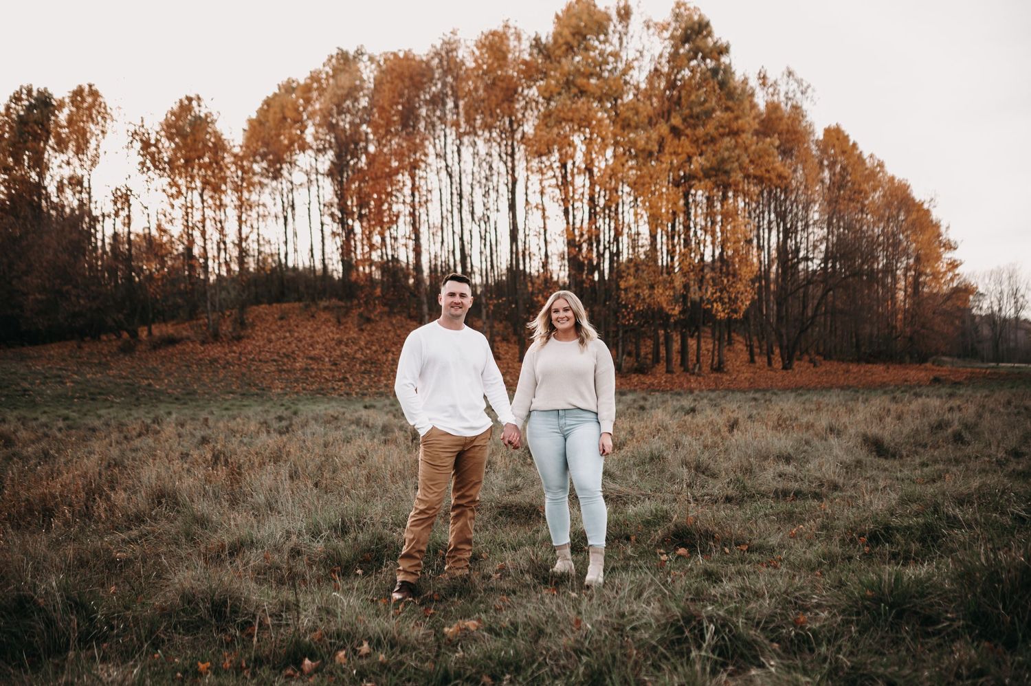 Couple holding hands while standing in field with orange autumn trees behind them.
