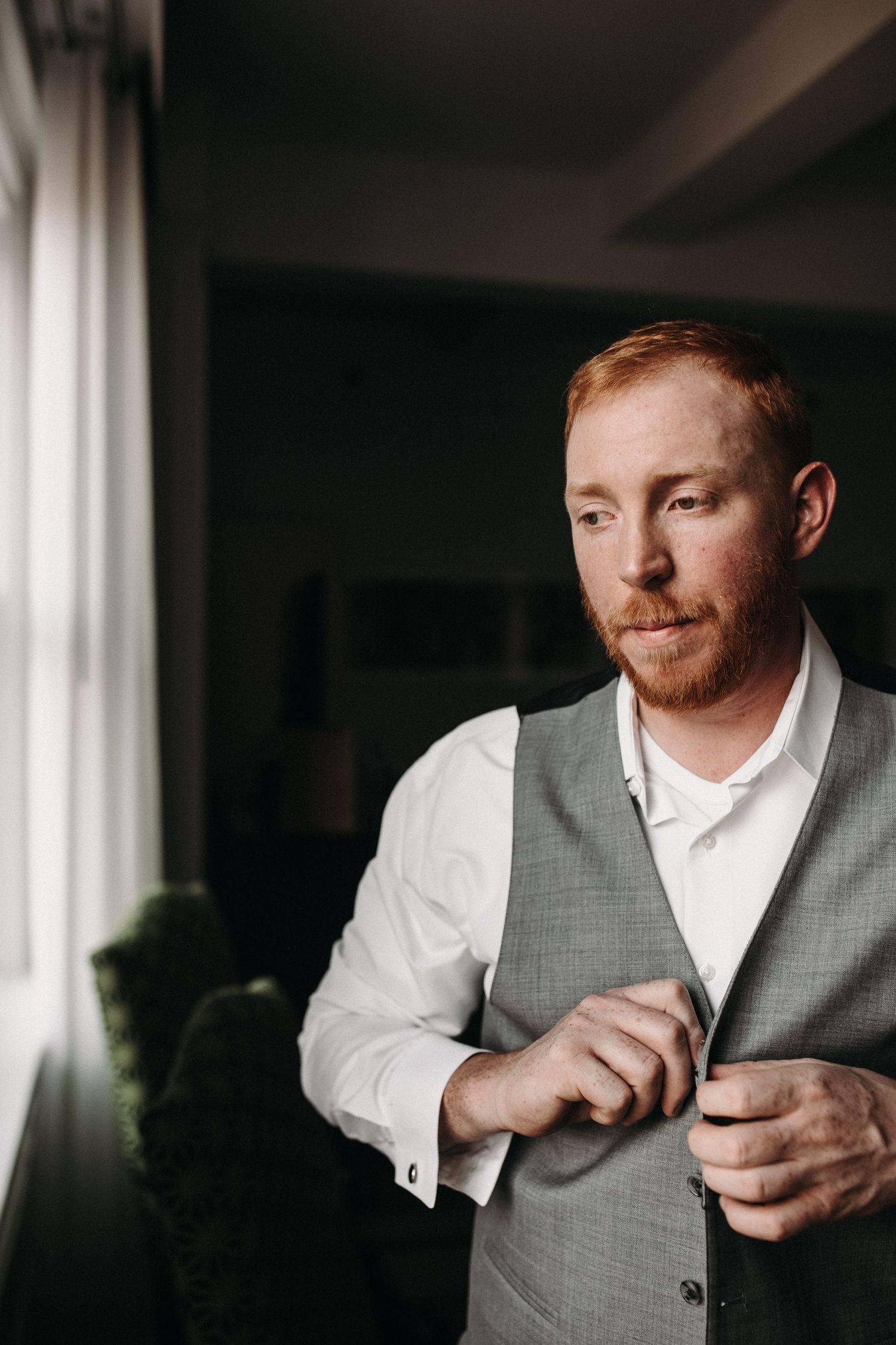 A person in formal attire buttons their grey vest while standing by a window.