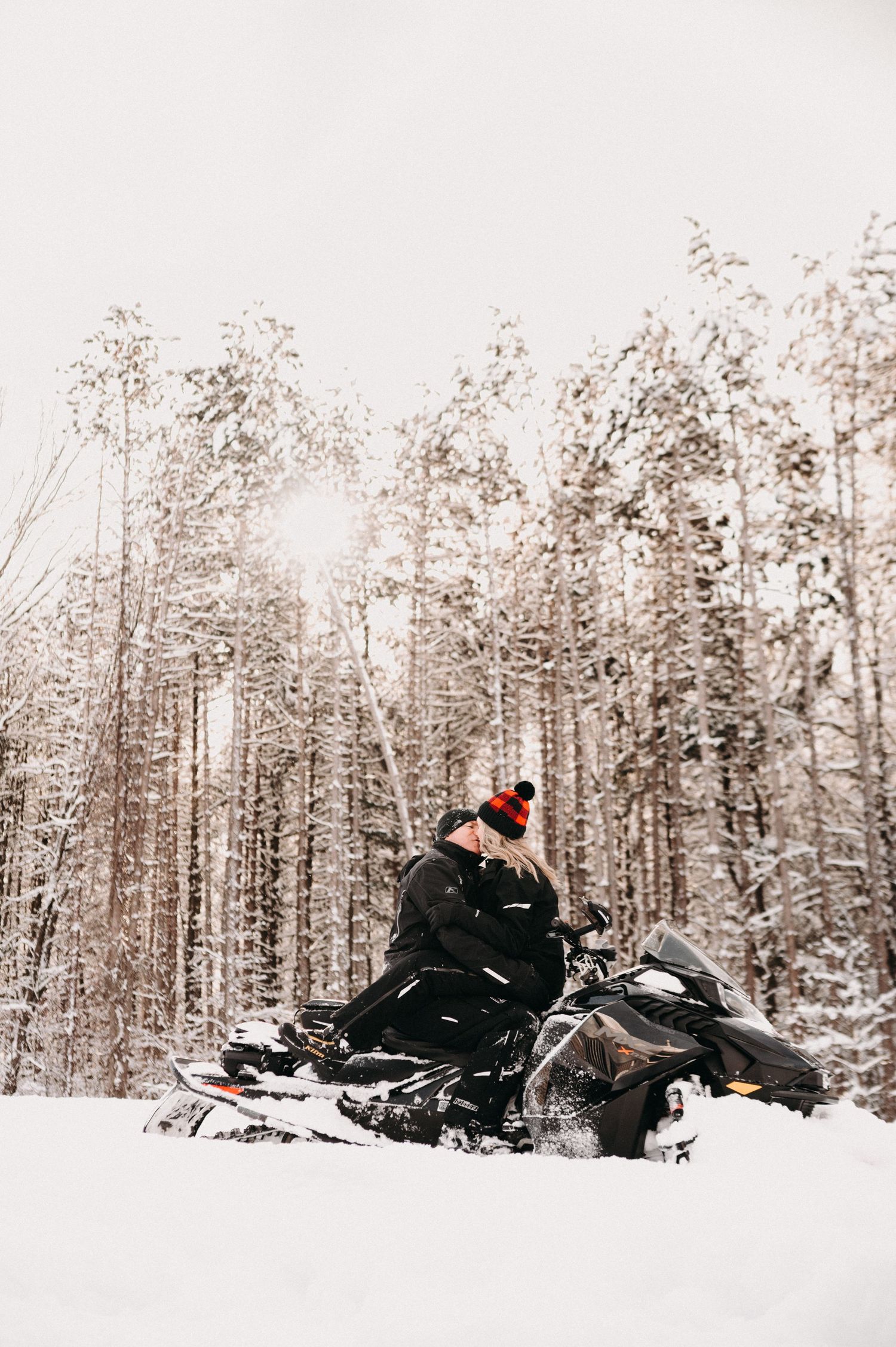 A snowmobiler in black winter gear poses against a backdrop of snow-covered trees.