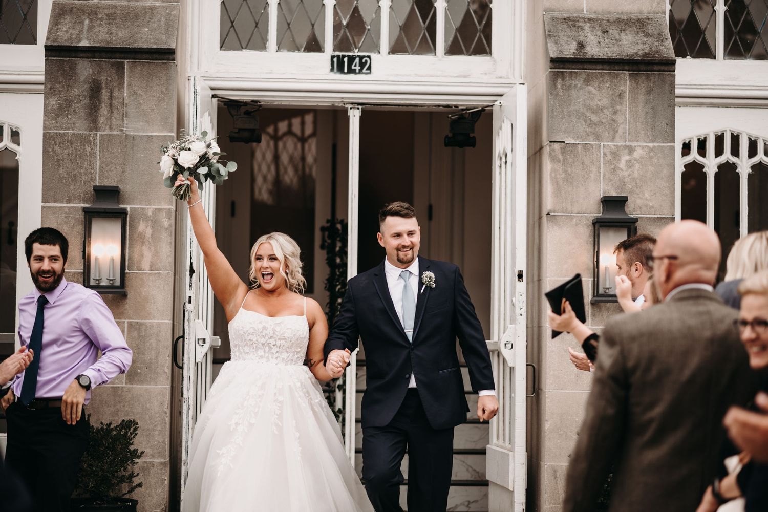 A newlywed couple exits their wedding ceremony with raised hands in celebration.