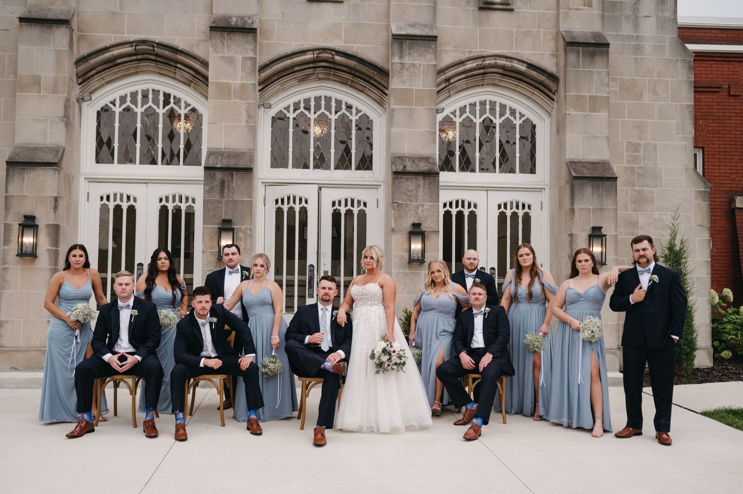 Wedding group poses casually in front of stone church with Gothic windows.