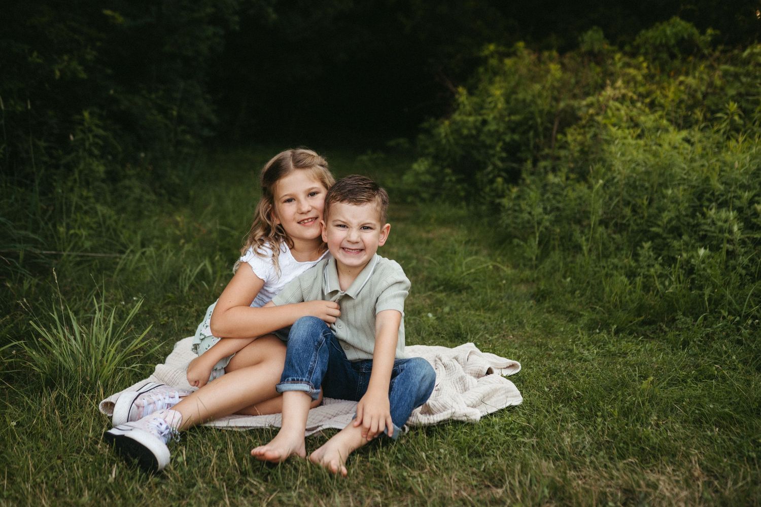 Two children sit close together on a white blanket in a grassy outdoor setting.