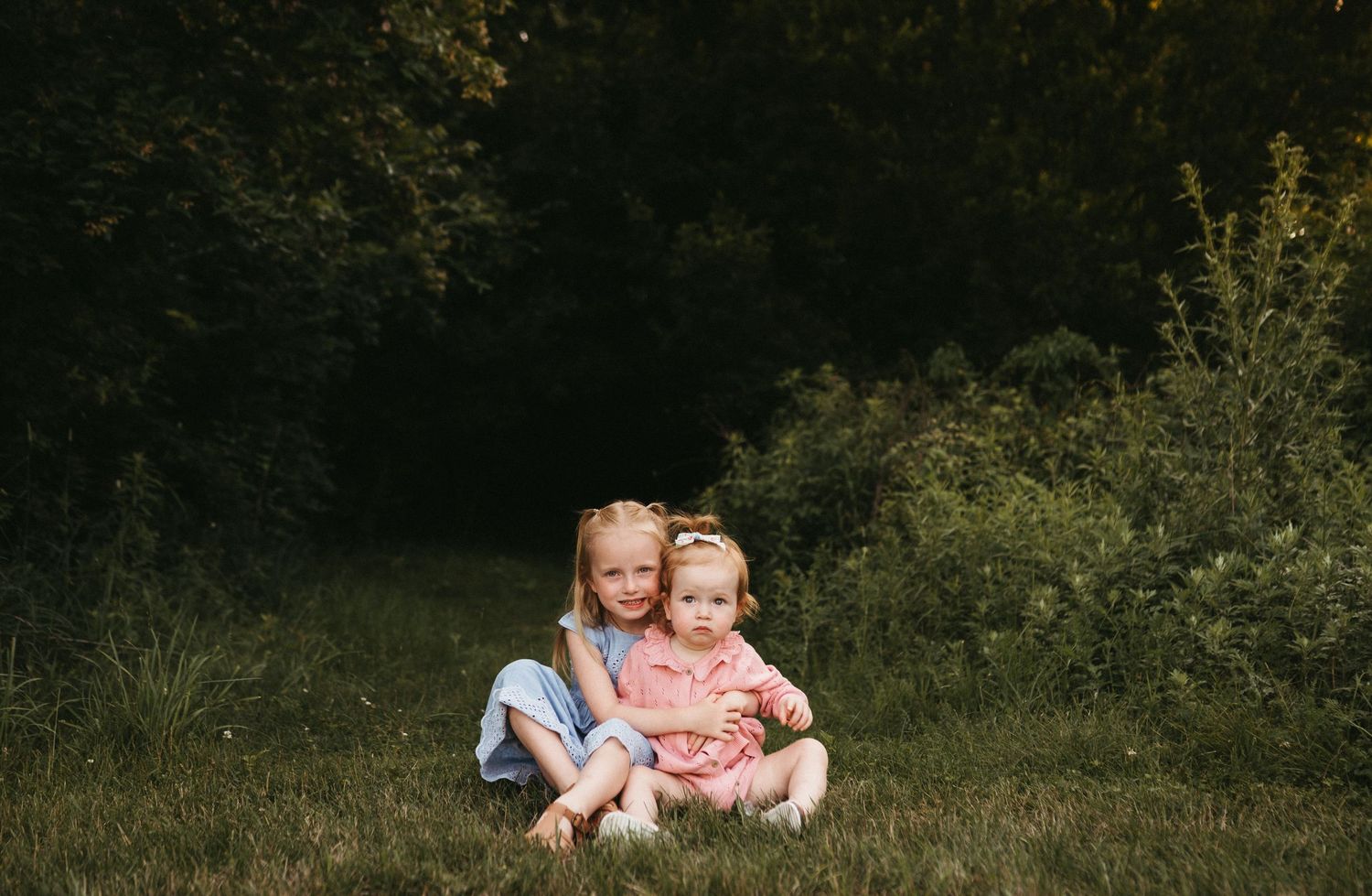 Two young children sit together on grassy ground with green foliage in the background.