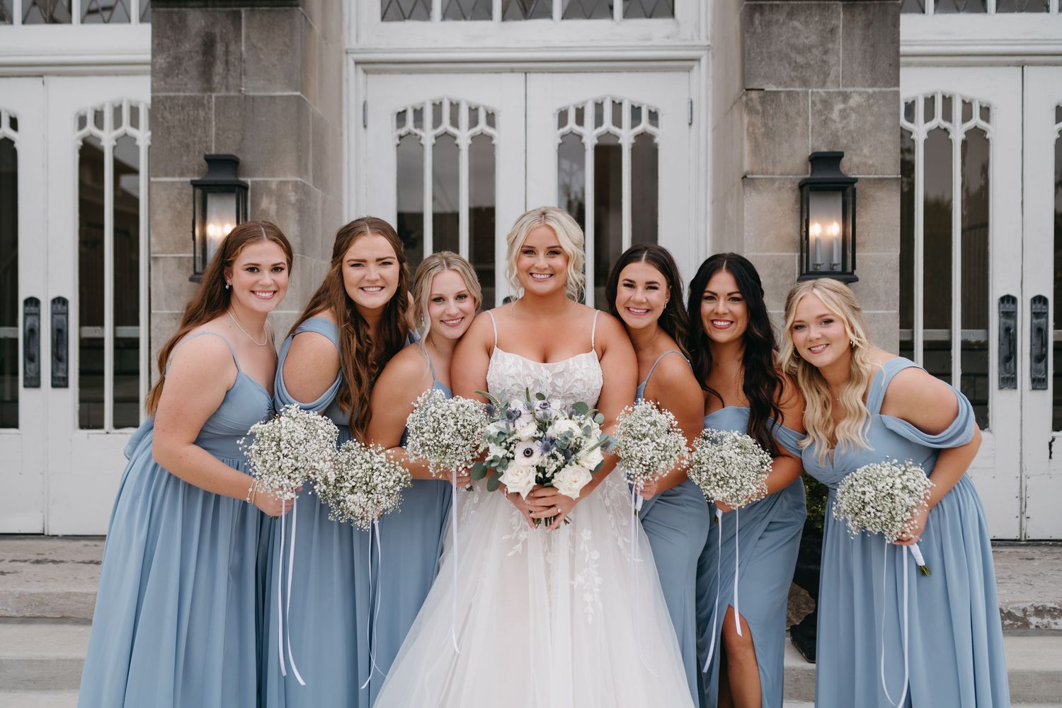 Bridal party in light blue dresses holds baby's breath bouquets against gothic church backdrop.