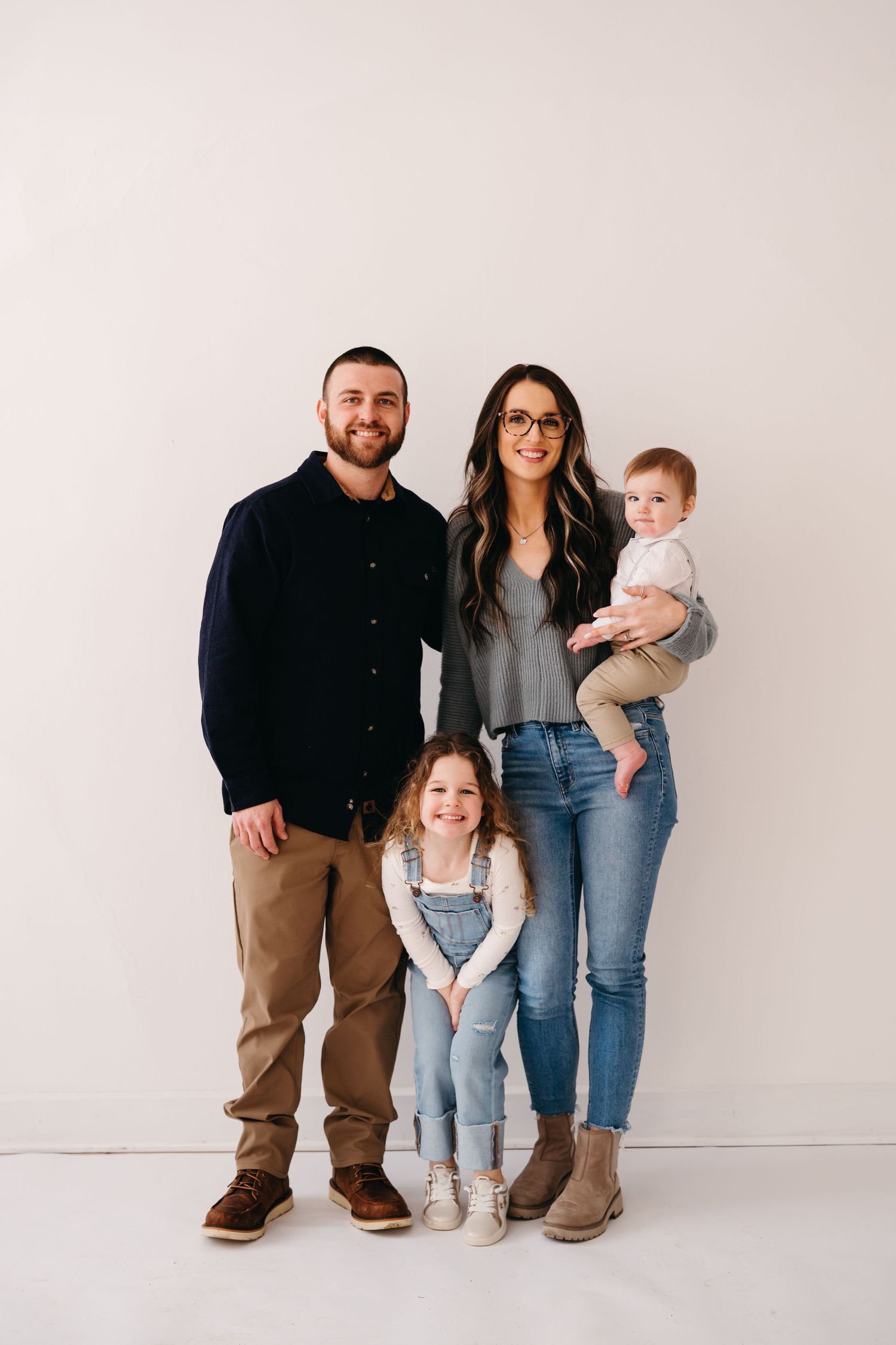 A family wearing casual outfits stands together against a light colored backdrop for a minimalist portrait.