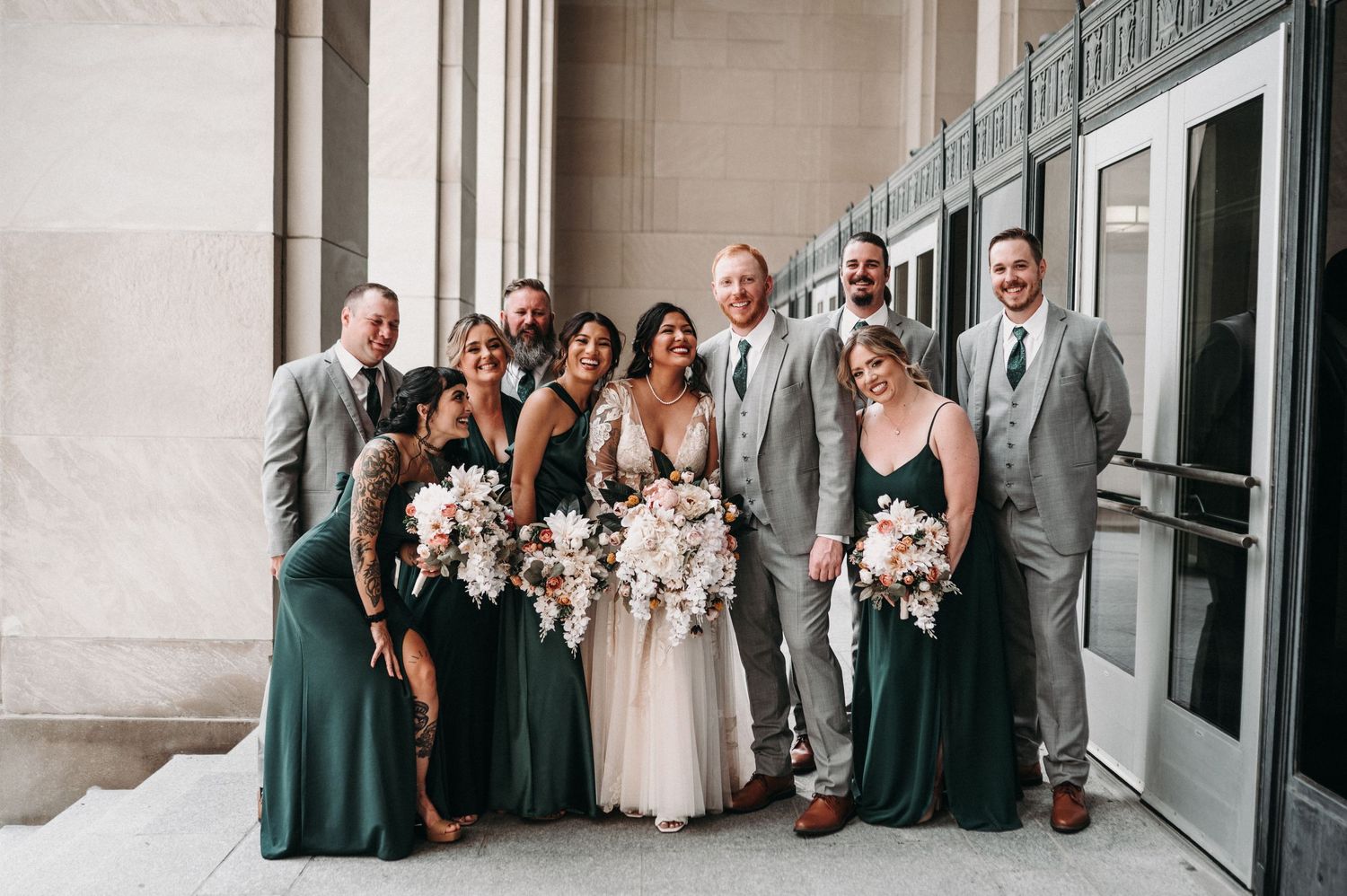 Wedding party in emerald green dresses and gray suits poses together in grand hallway.