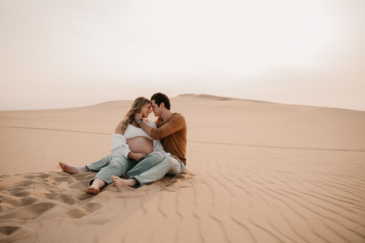 Couple sits together intimately on desert sand dunes during golden hour sunset.