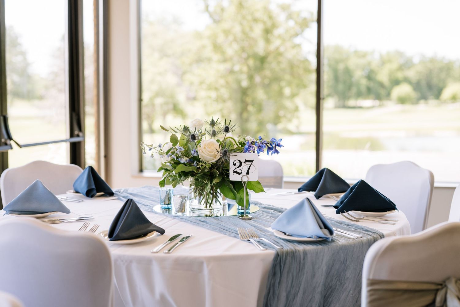 Elegant wedding reception table setting with blue napkins and white linens in a bright venue with large windows.