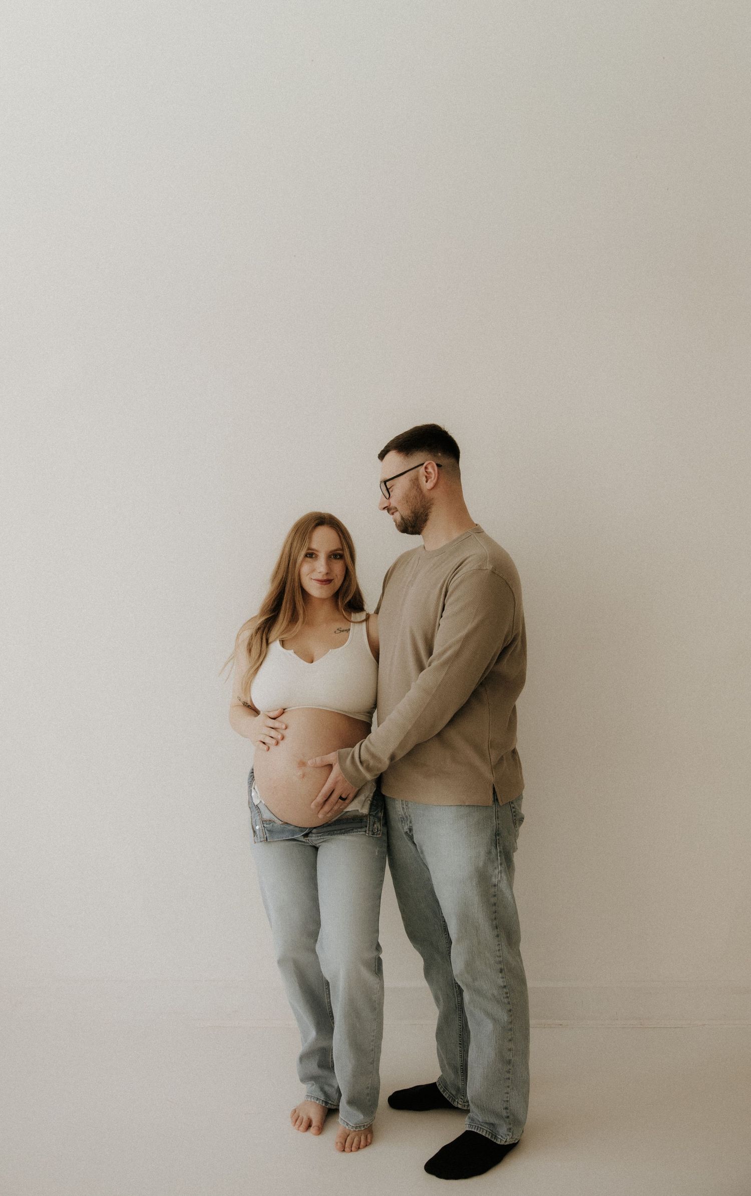Intimate maternity photoshoot with couple wearing casual attire against neutral background.