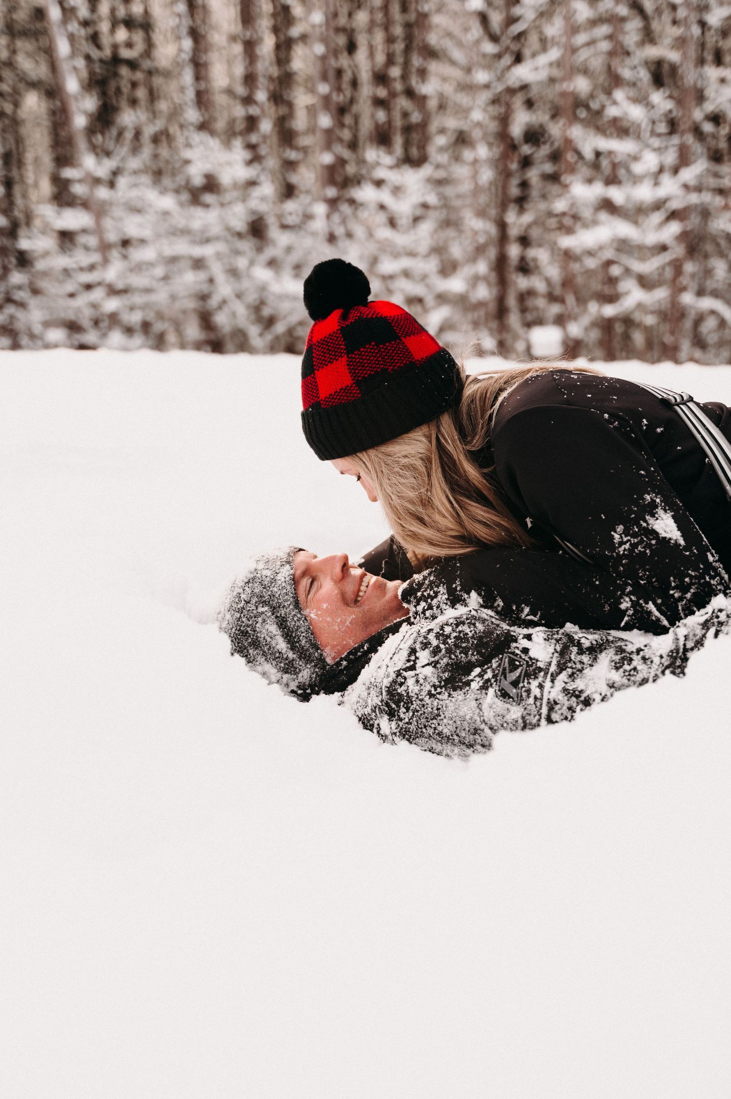 Couple in winter attire playing in deep snow with forest background.