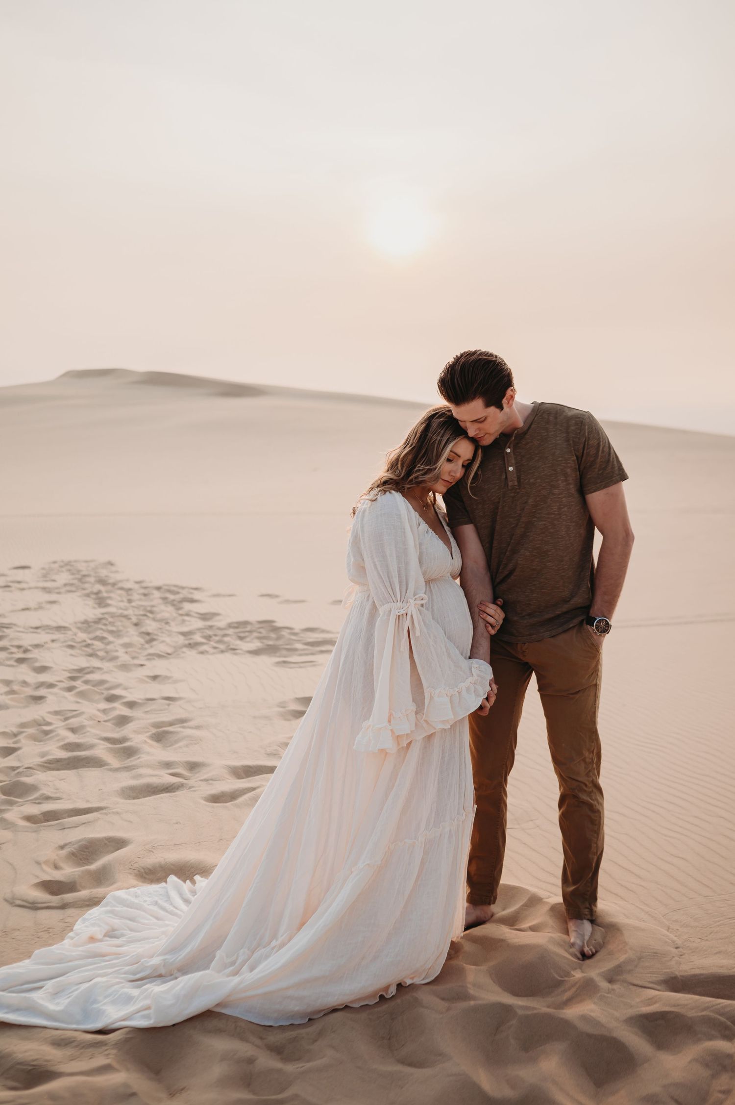 A romantic couple shares an intimate moment at sunset in the desert sand dunes, with her flowing white dress trailing behind.