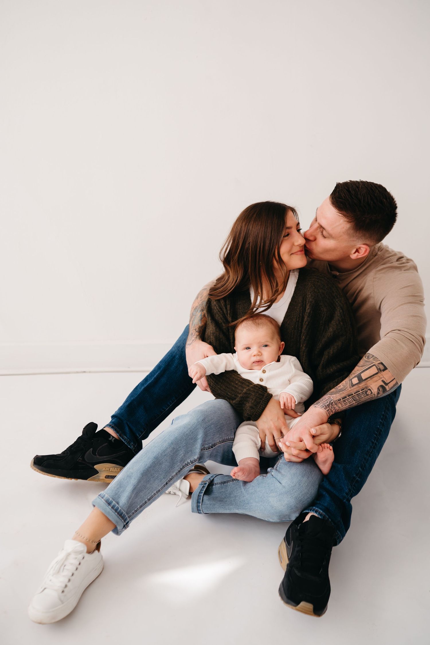 A young family sits together on a white floor as a couple shares a tender moment while holding their baby.