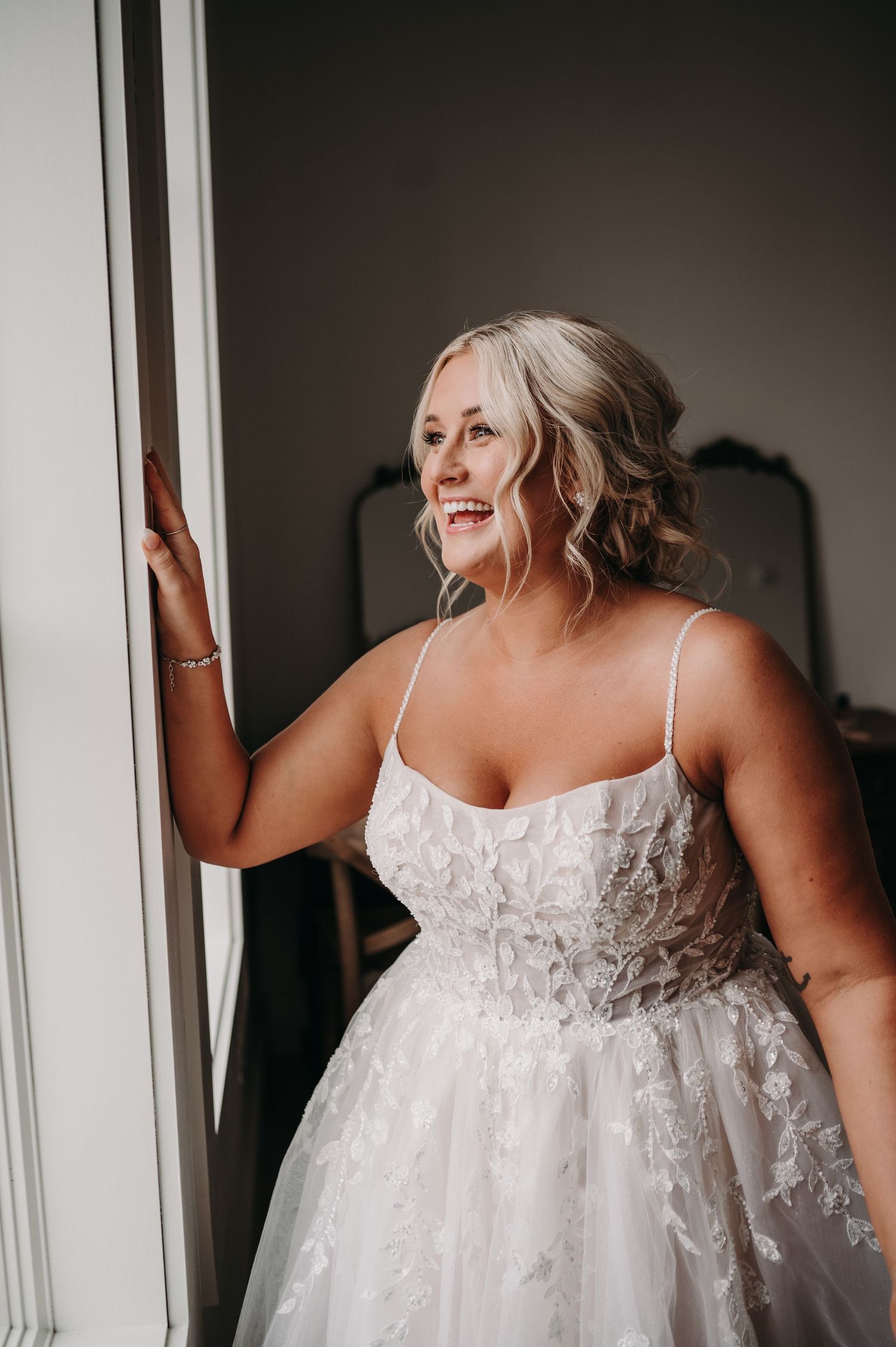 Joyful bride in lace wedding gown poses by window in natural light.