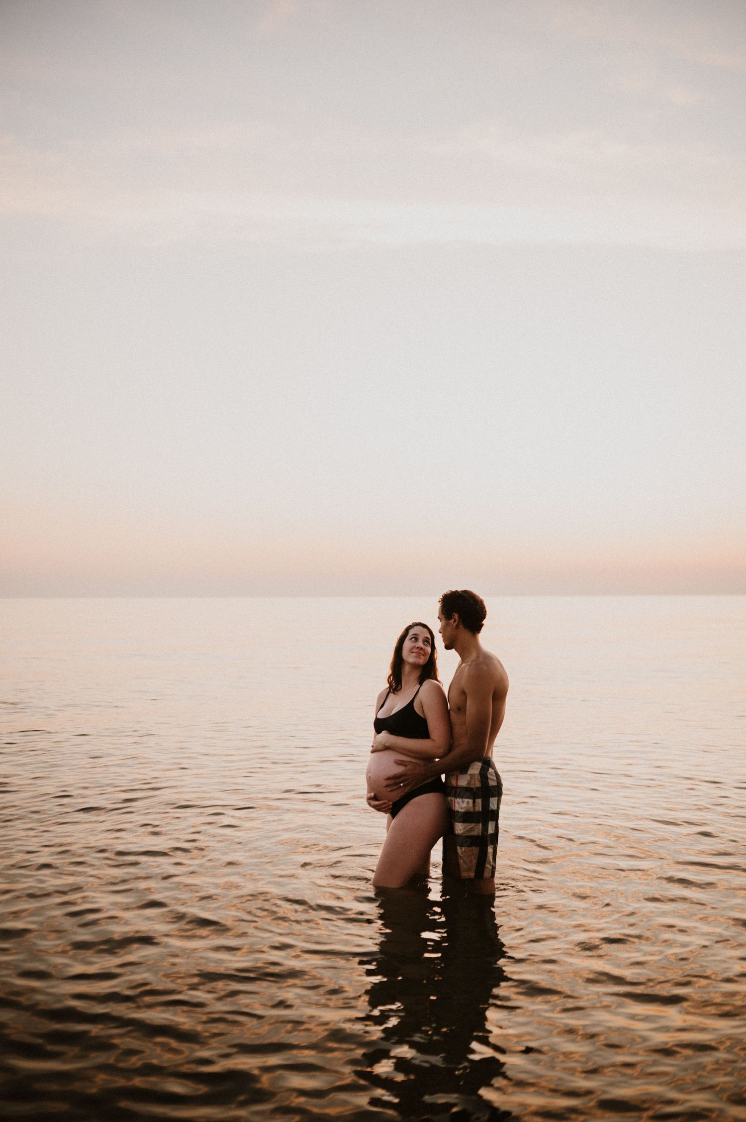Romantic maternity photo of couple embracing in calm ocean waters during golden hour.