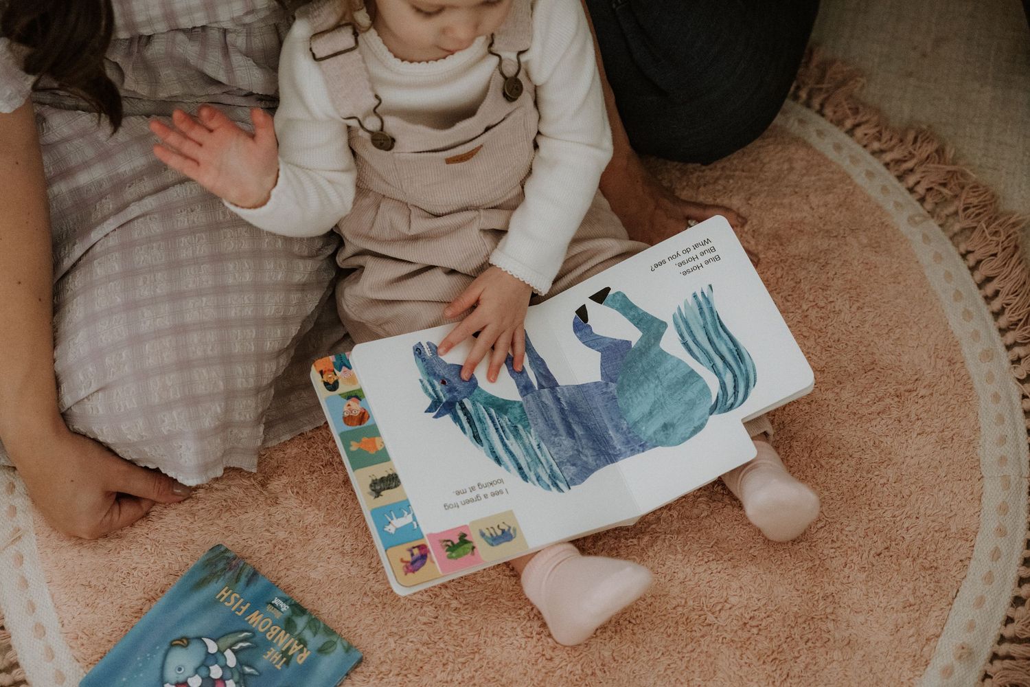 Children sitting on beige carpet looking at books and toys spread out on the floor.