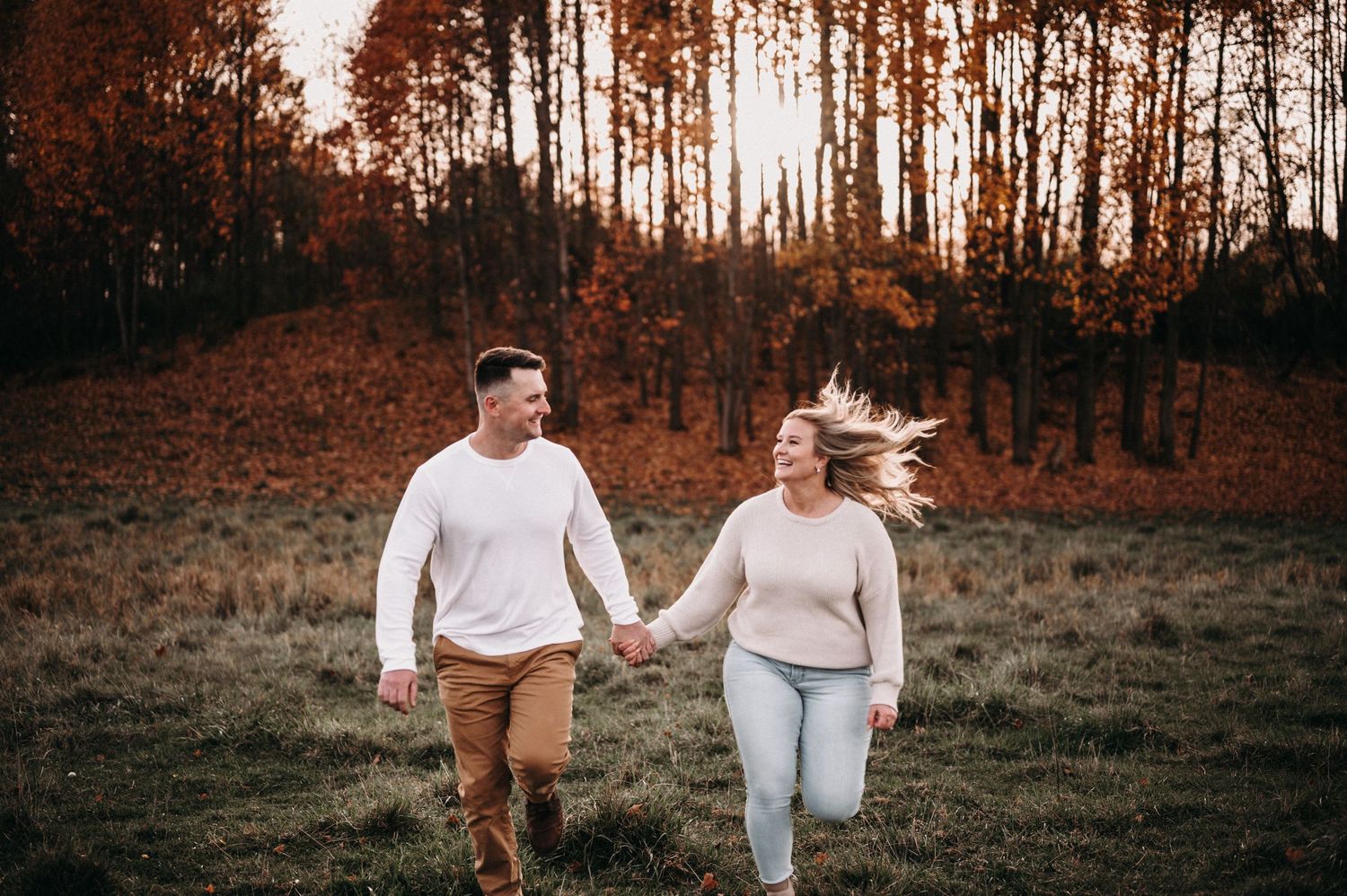 Couple holding hands while running through an autumn field with orange trees in the background.