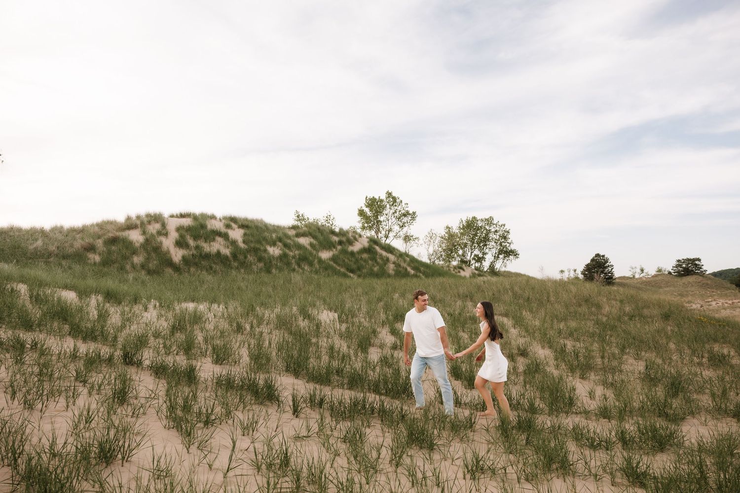 Couple holding hands walking through sandy dunes and green grass under a cloudy sky.