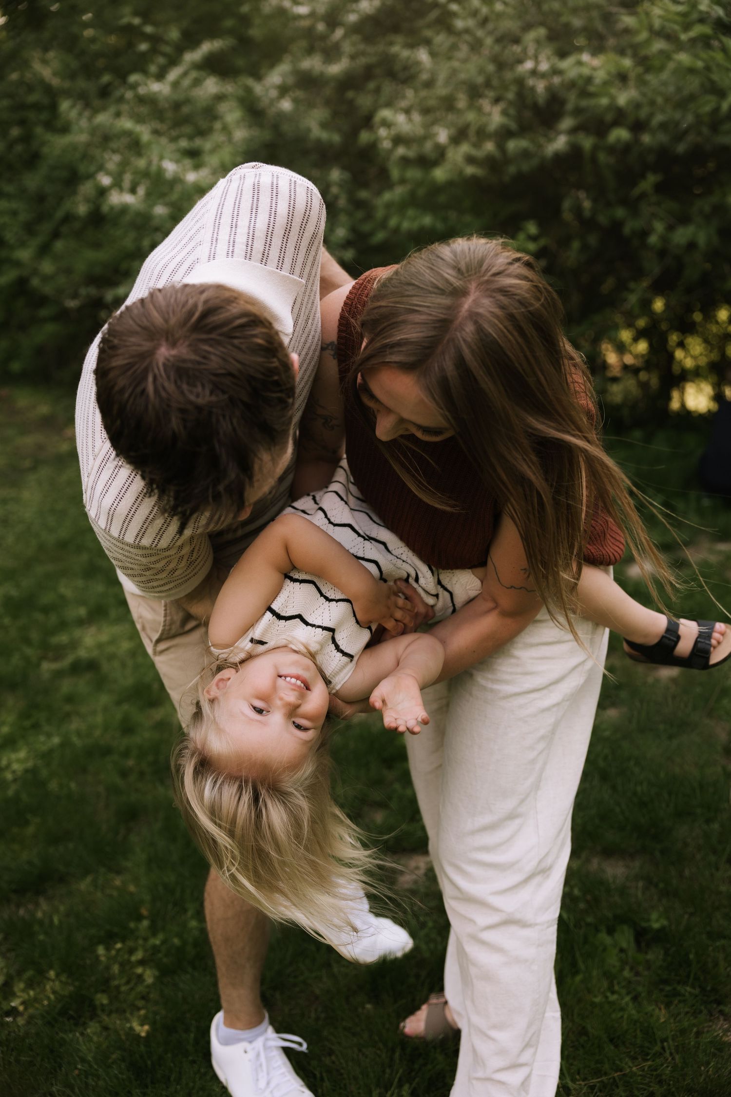 Parents playfully flip their laughing toddler upside down in a lush green backyard.
