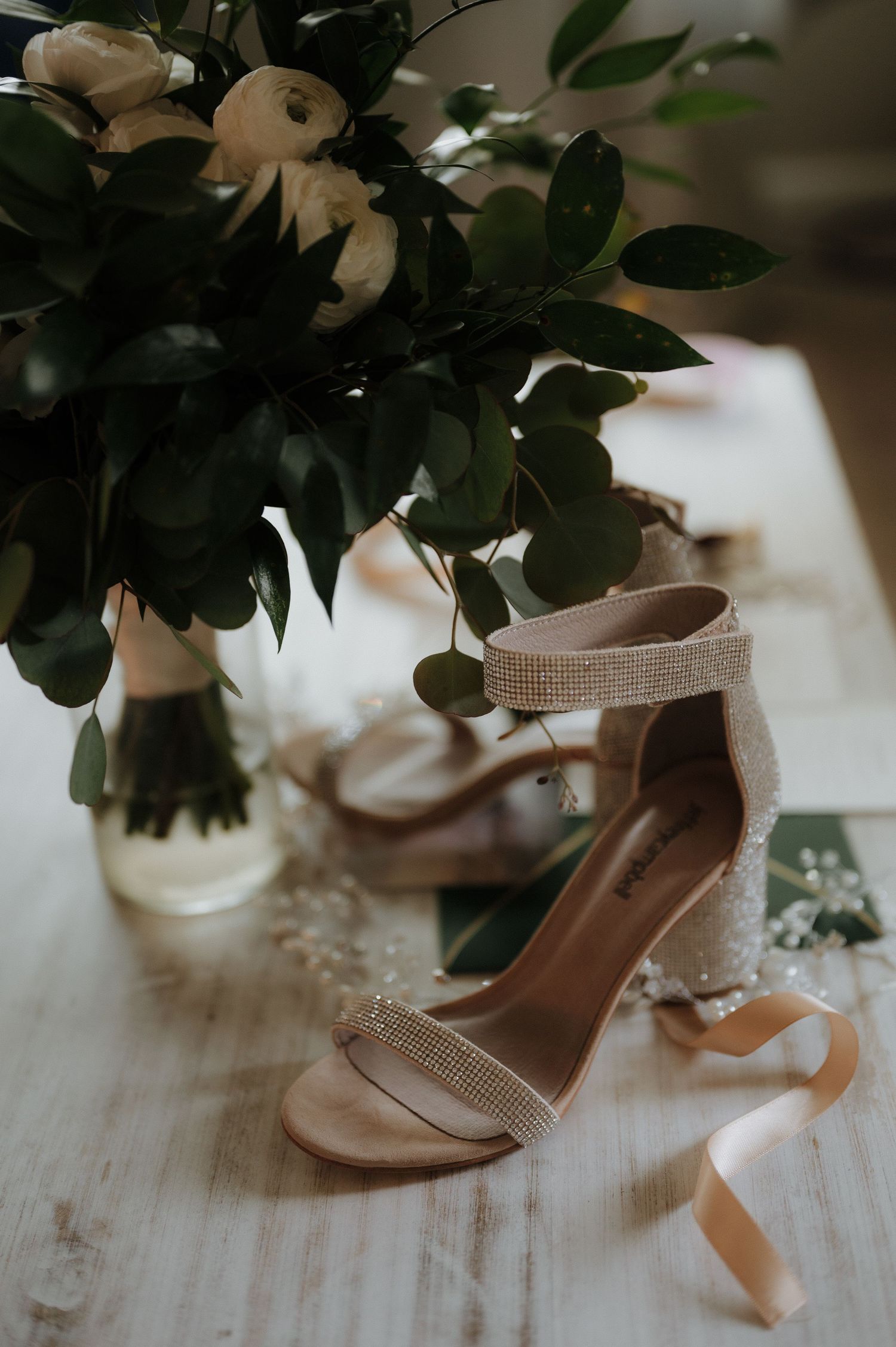 Elegant rhinestone block heel bridal sandals displayed on a white table beside a white floral bouquet with greenery.