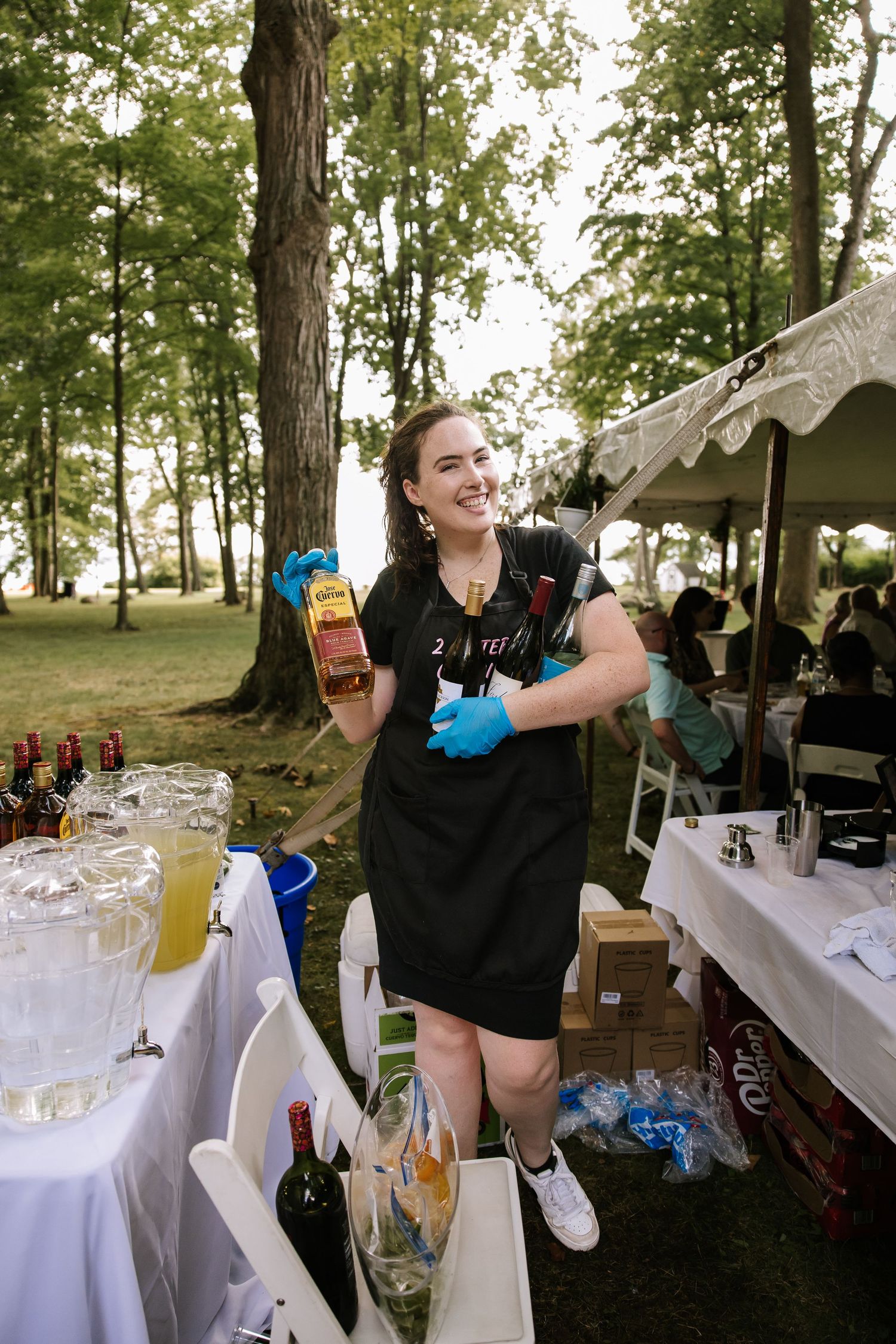 Wedding reception setup with drinks being arranged on white-clothed tables outdoors.
