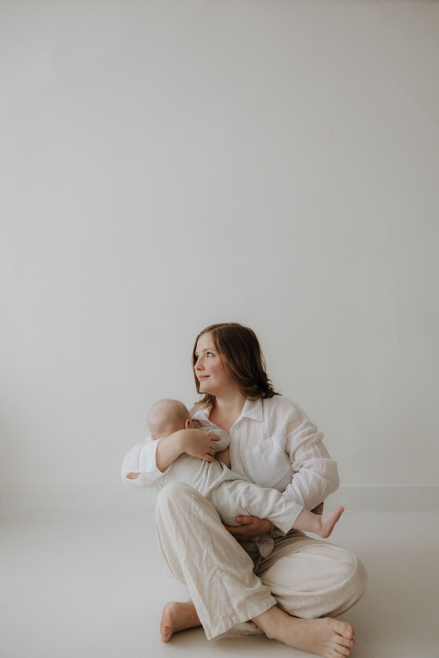 Mother sitting cross-legged on white floor holding her newborn baby lovingly.