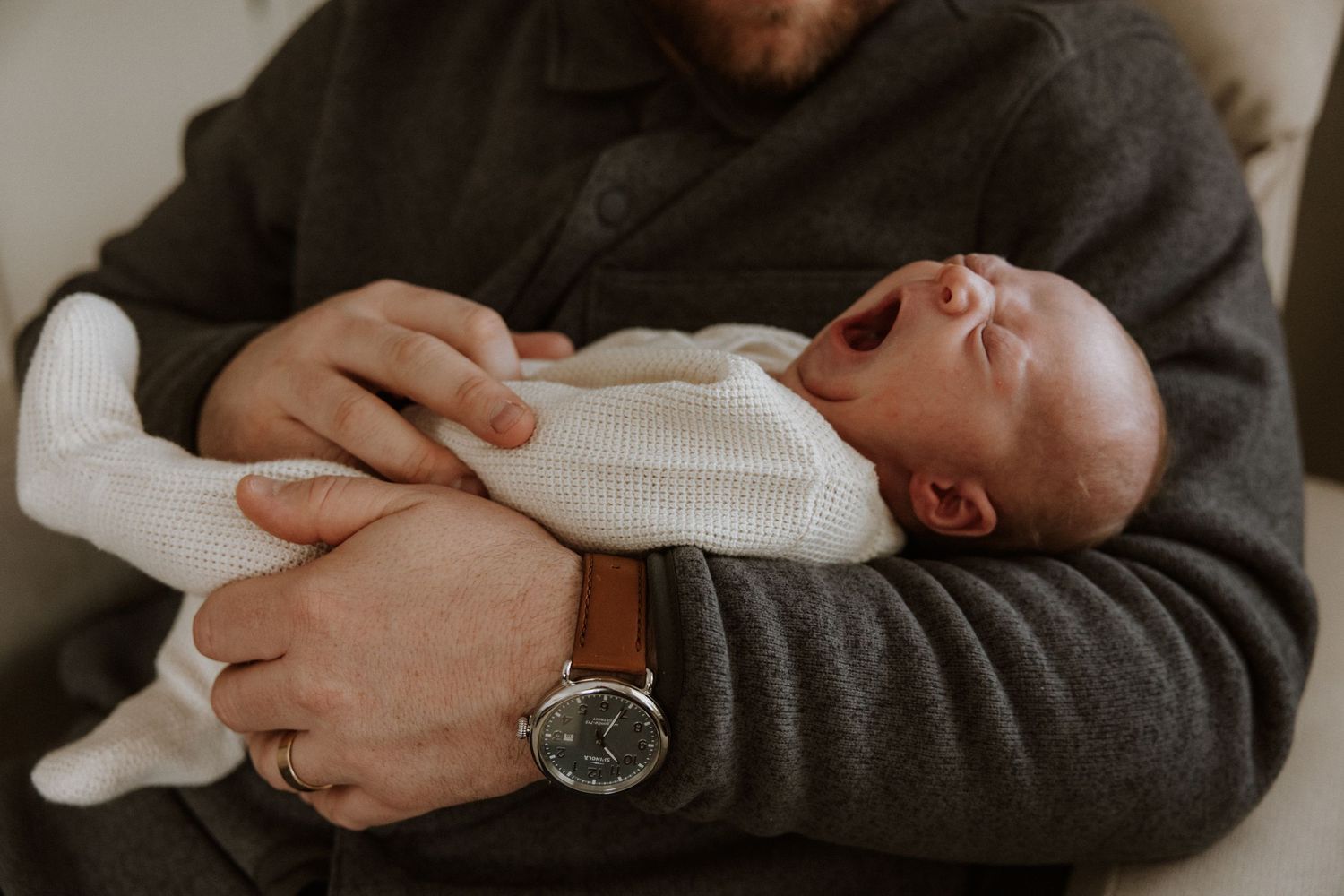 A sleeping infant in white clothing cradled in arms wearing a gray sweater.