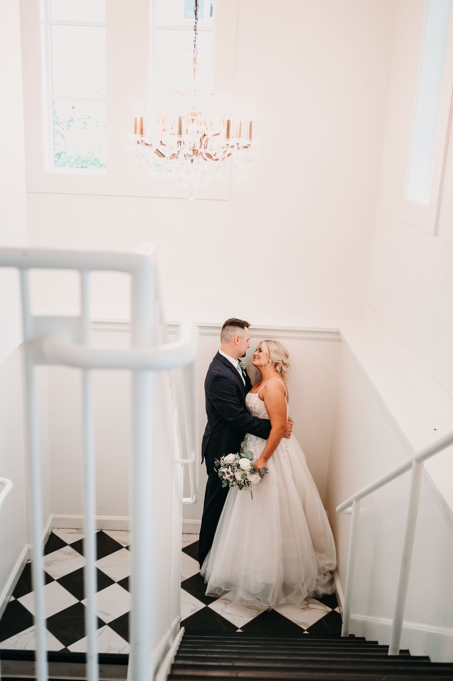 Intimate wedding photo on a staircase with a white railing and chandelier overhead.