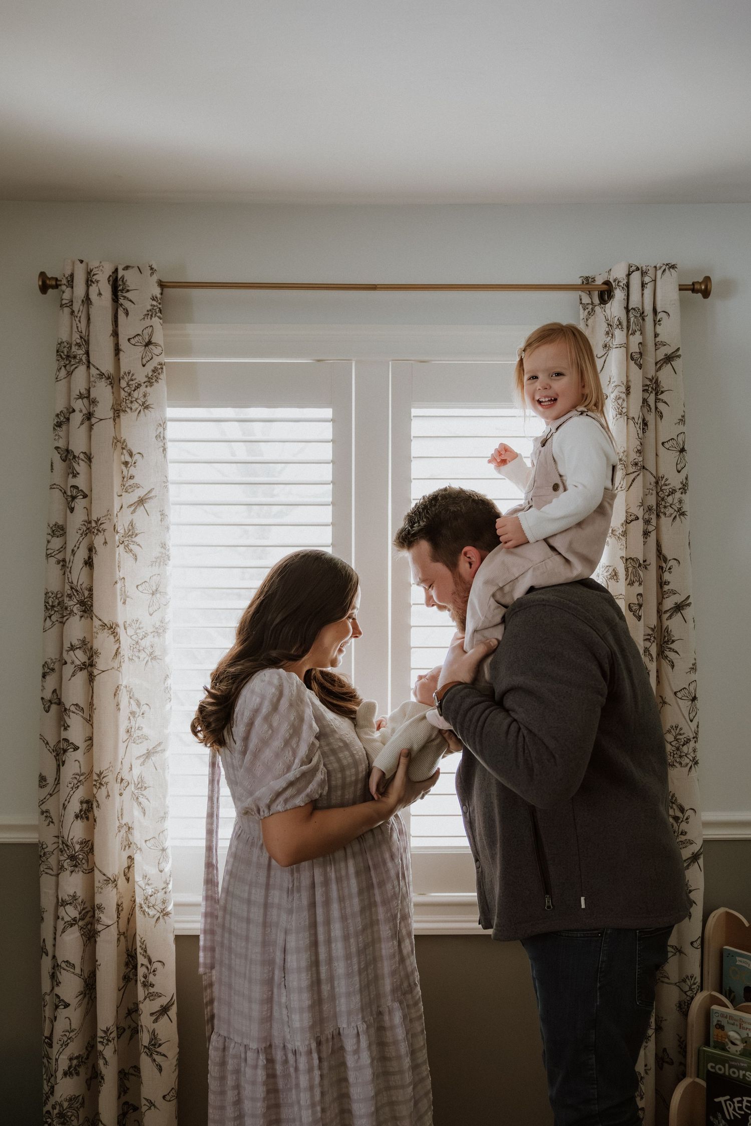 A family shares a joyful moment by a window with floral curtains in a sunlit room.
