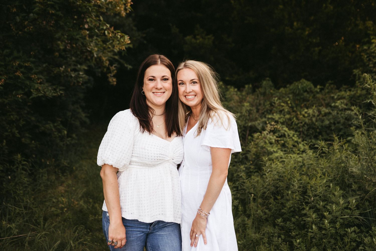 Two women smile together outdoors wearing white tops against a natural background.