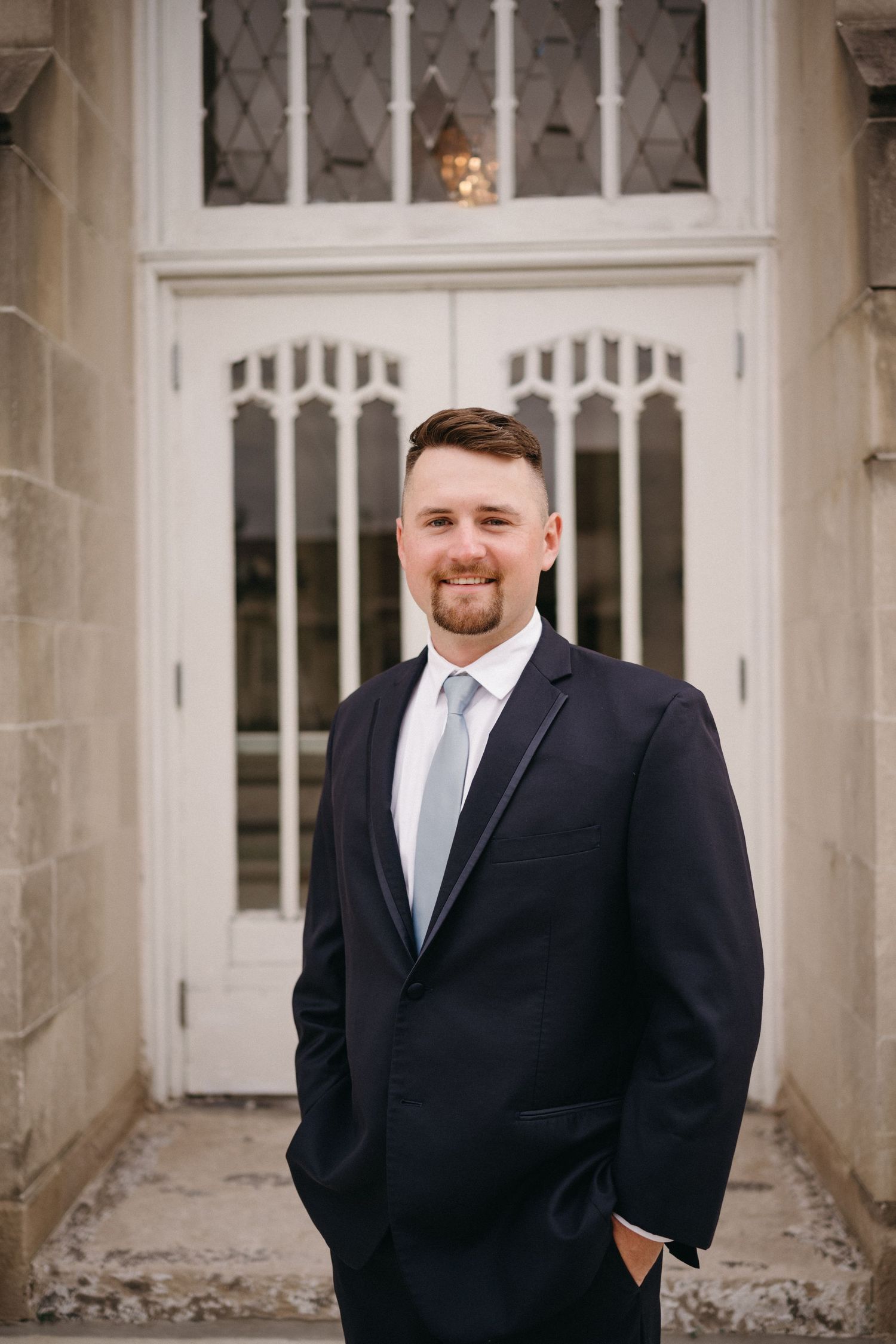 Groom poses in classic navy suit in front of gothic-style church doors.