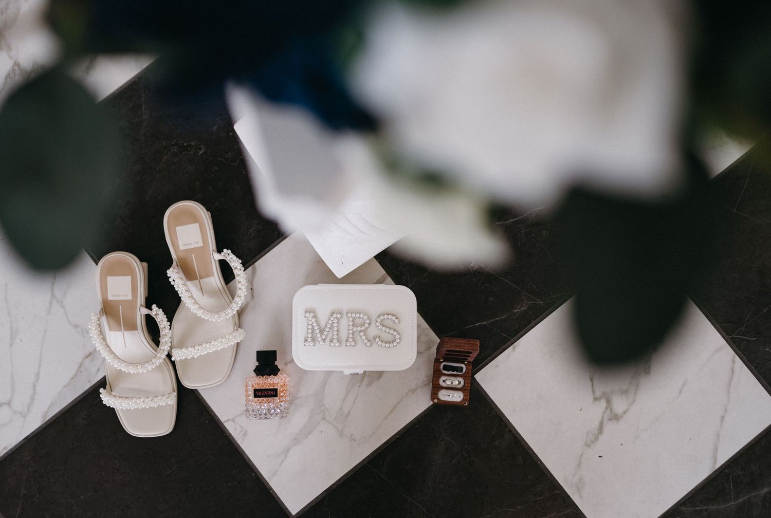 Wedding accessories including white sandals and a 'MRS' clutch displayed on marble.