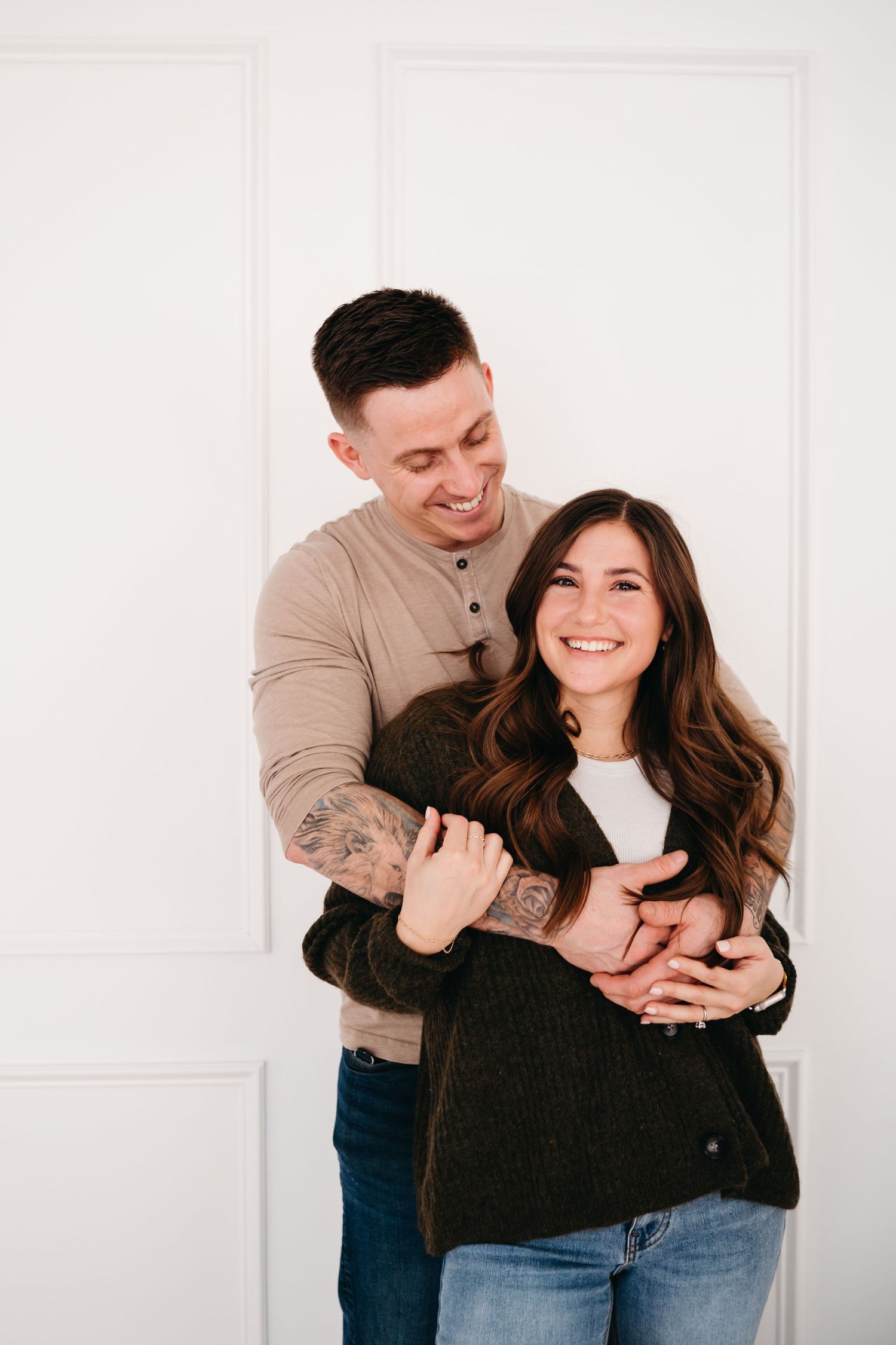 Couple embracing and laughing together against white wall in casual attire.