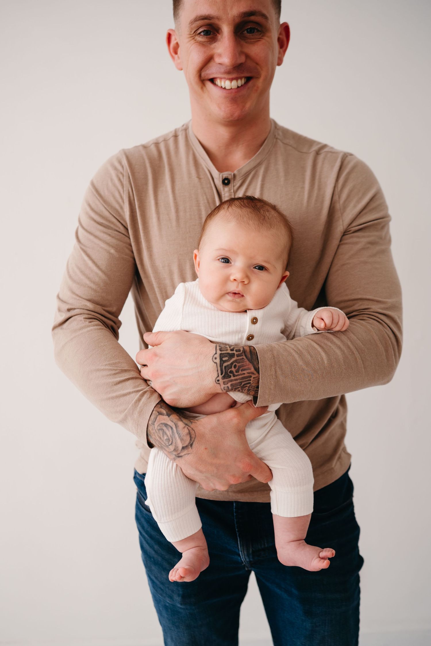 Parent wearing beige henley holds infant dressed in white against minimalist backdrop.