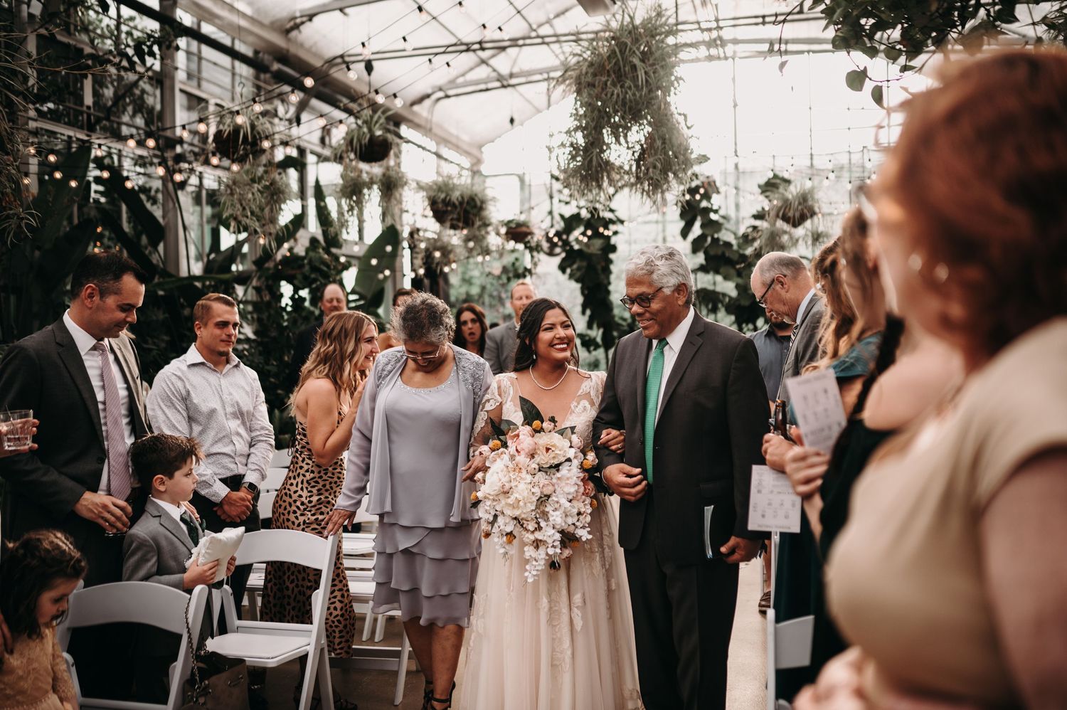 Wedding ceremony taking place in a sunlit greenhouse with tropical foliage and white chairs.