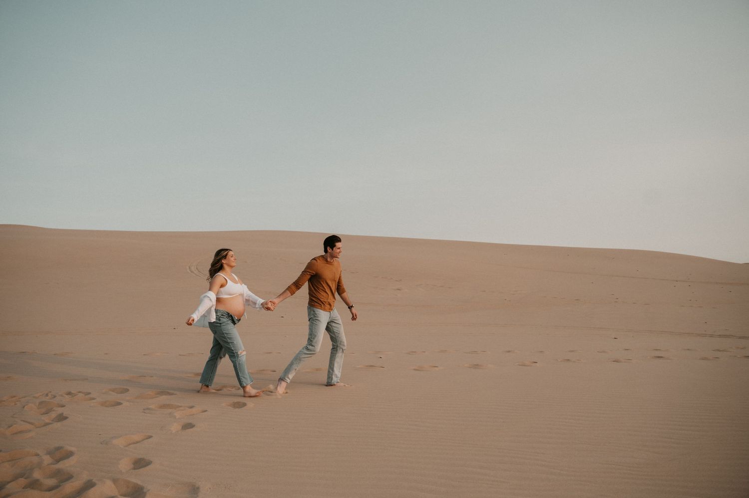 Couple walks hand in hand across desert sand dunes during sunset photoshoot.