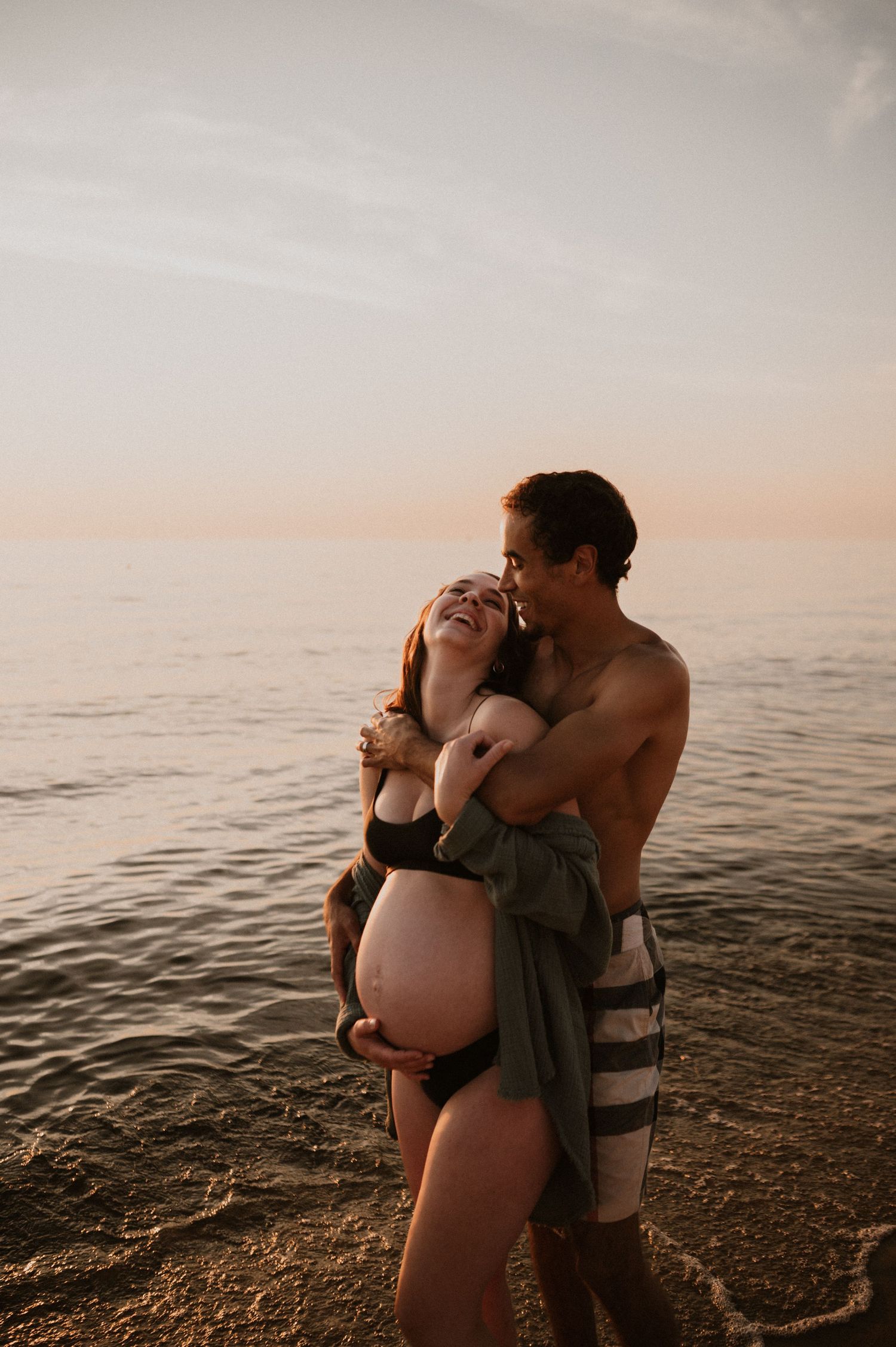 Candid maternity moment captured on beach during golden hour with ocean backdrop.