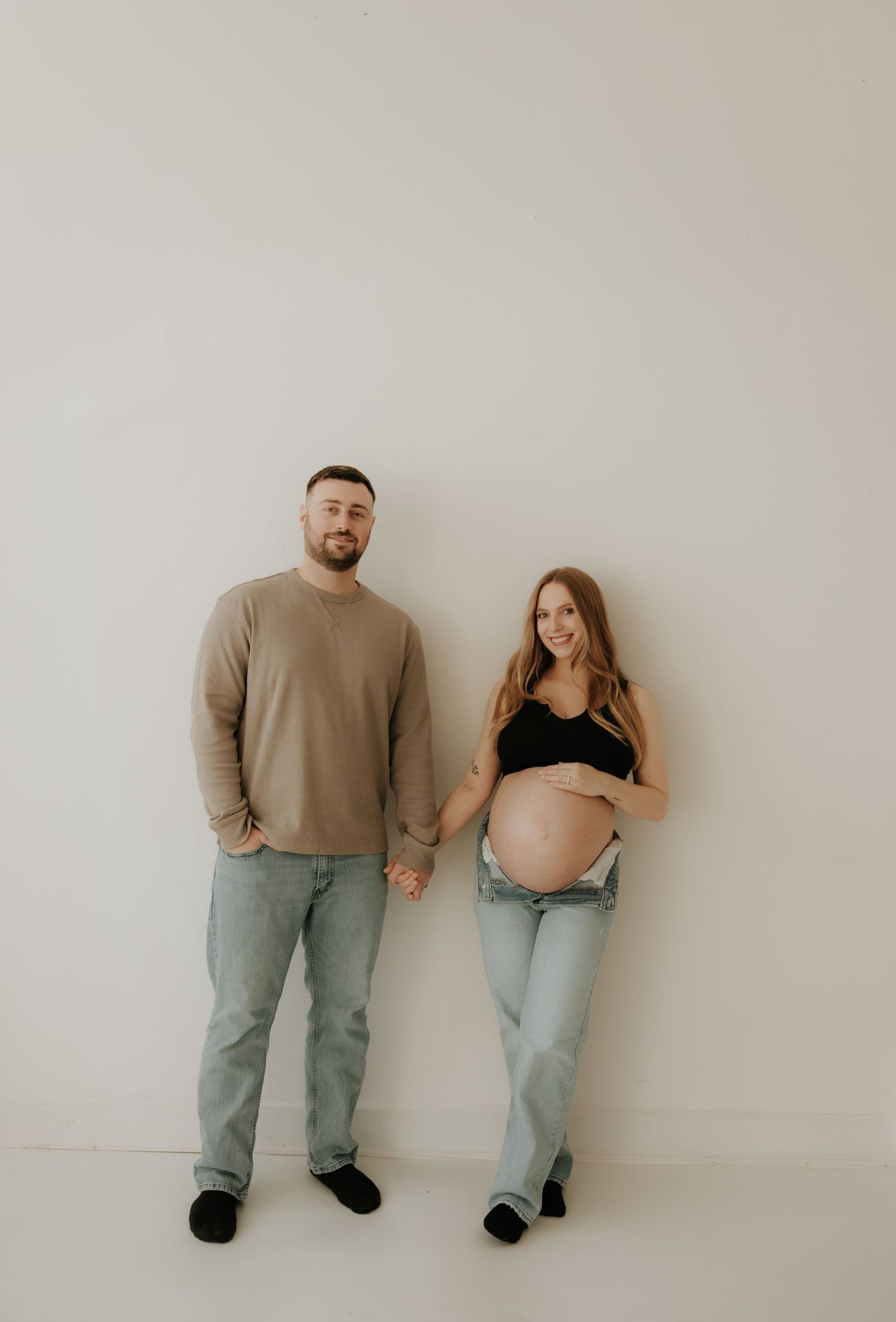 Candid maternity photos of couple sitting against white wall in matching denim.