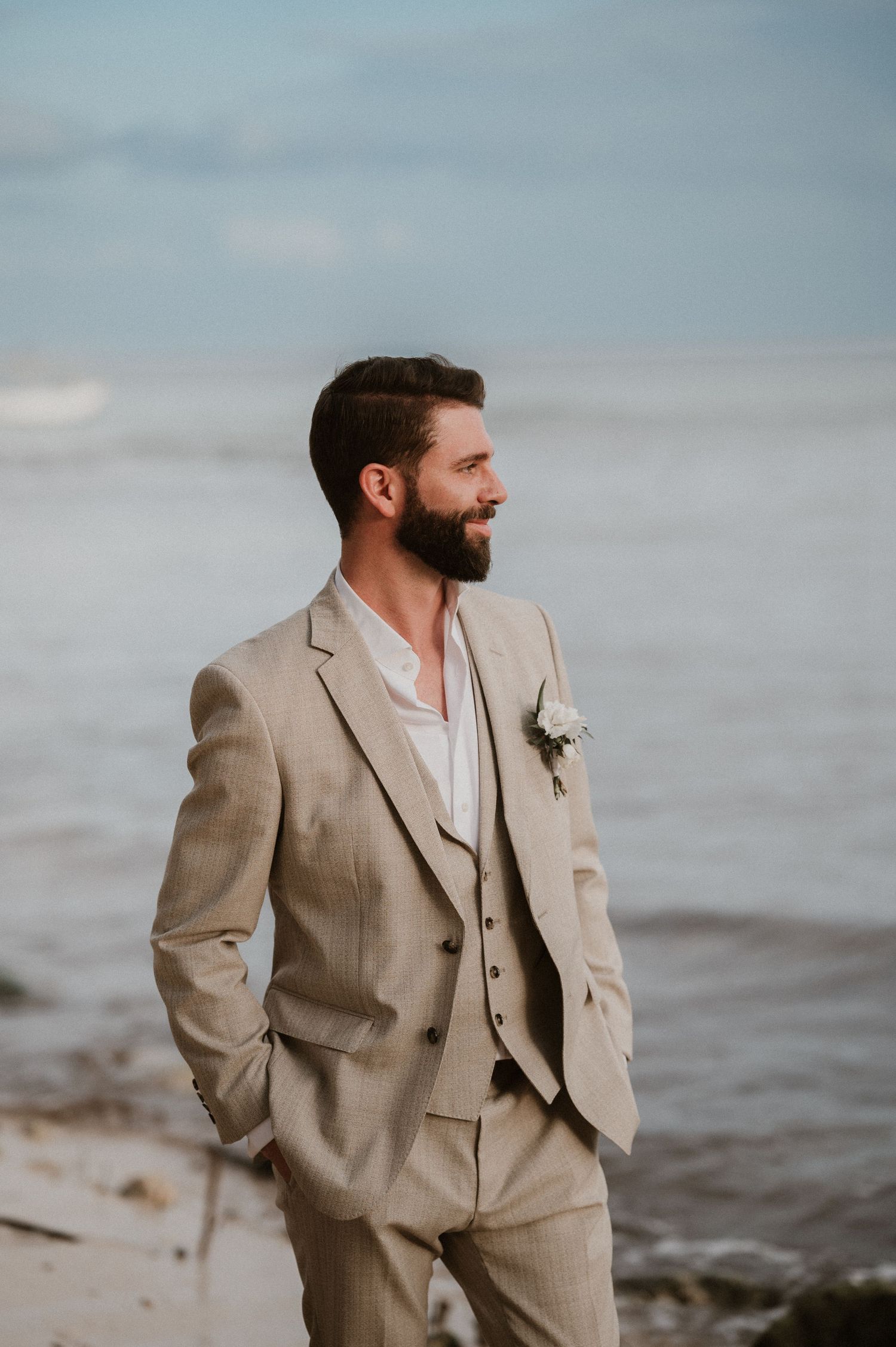 Groom in beige three-piece suit standing by ocean at sunset.