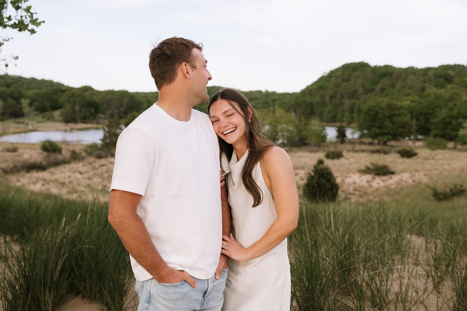 Happy couple in matching white outfits laughing together in a scenic outdoor setting with green hills and water.