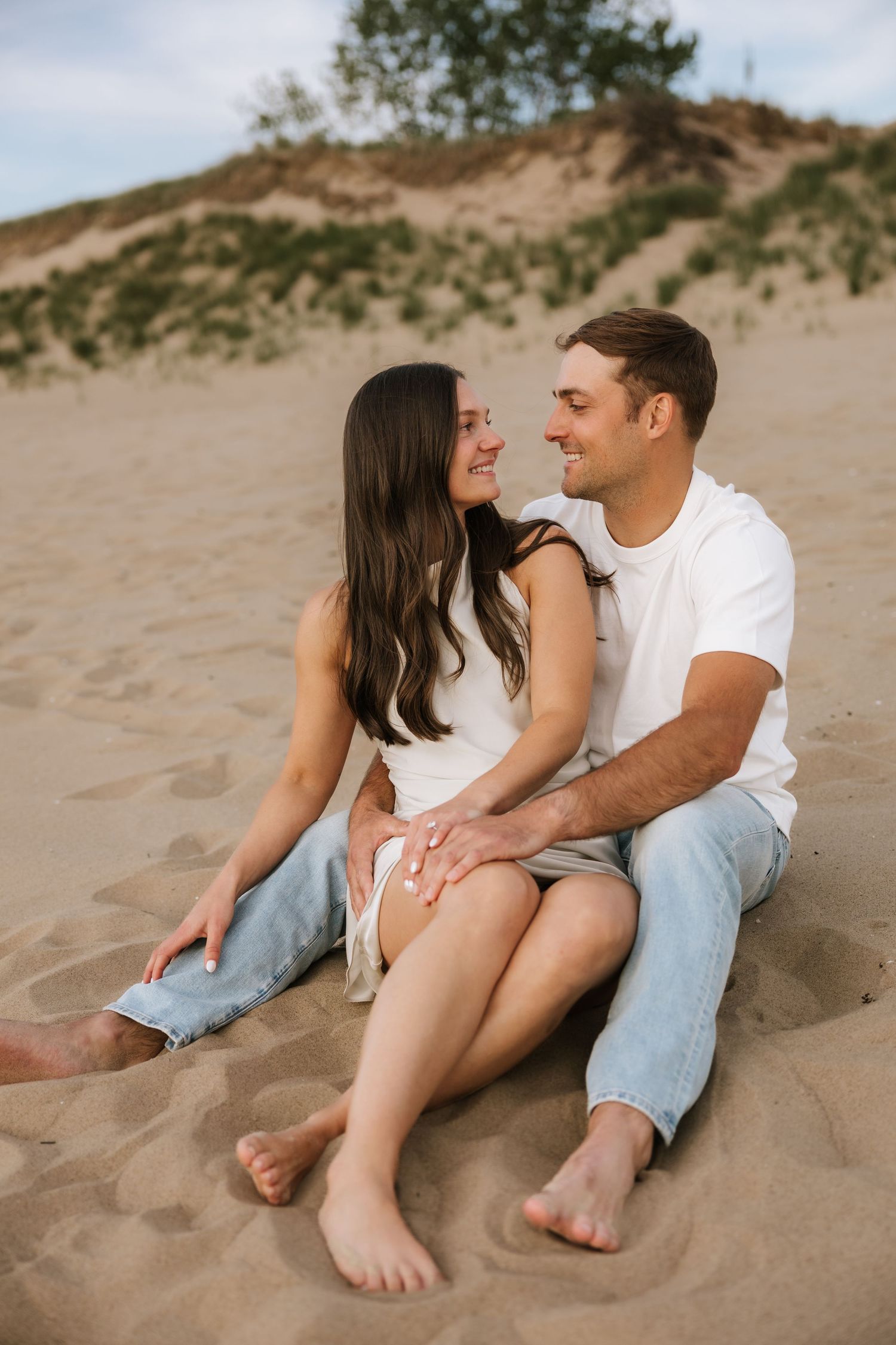 Couple sitting barefoot on sand dunes, smiling and gazing at each other affectionately.