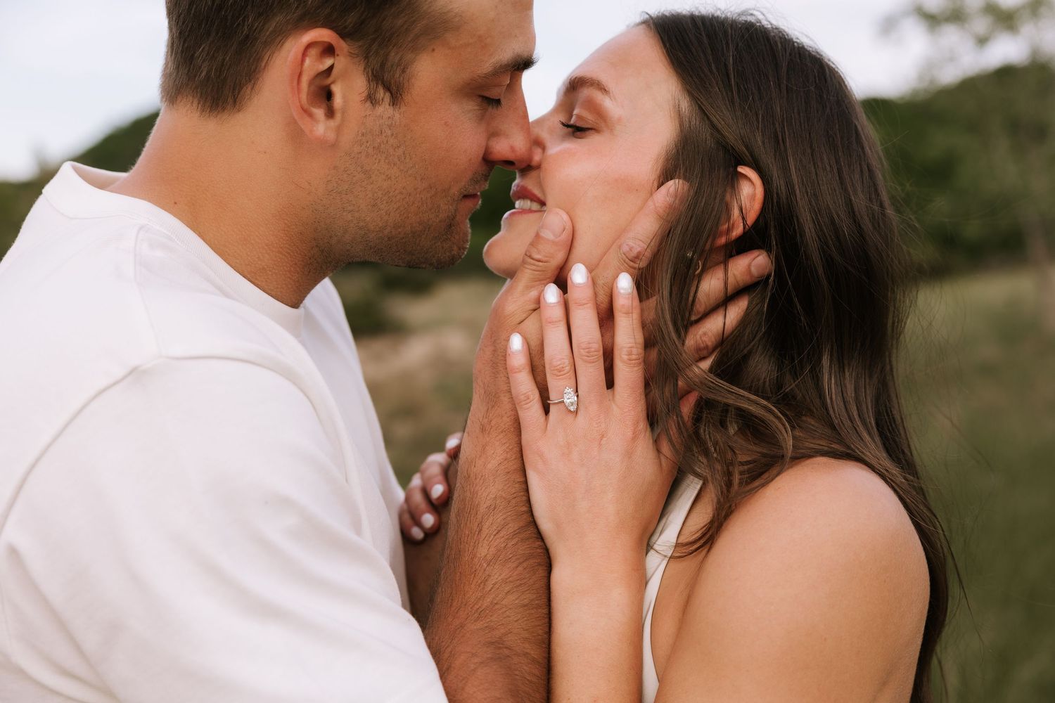 Engaged couple sharing a romantic kiss outdoors, diamond ring visible on her hand.